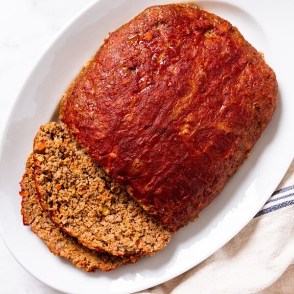 White platter sitting on white marble surface filled with meatloaf with two slices sliced off the side with ketchup all over the top.