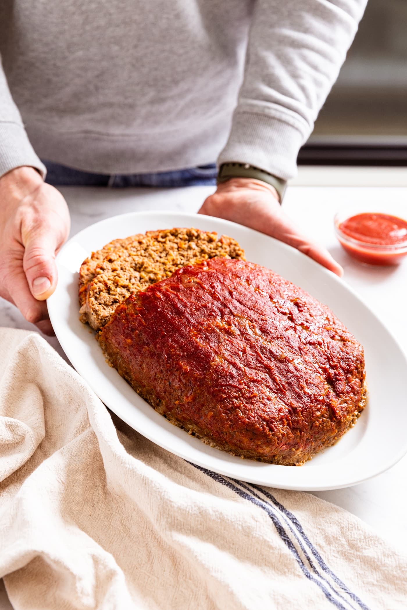 Hands holding white platter with meatloaf with two slices sliced off the back side with small bowl filled with ketchup to the side.