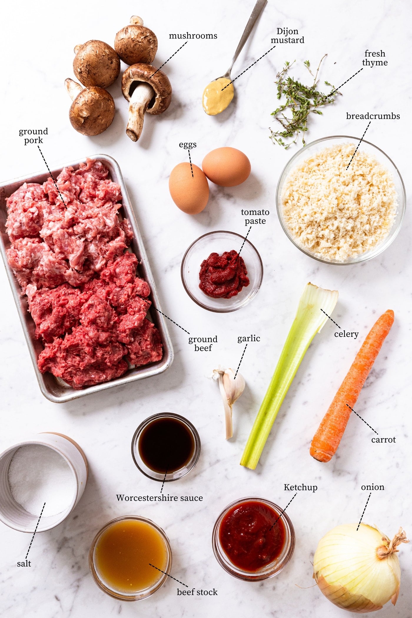 Marble surface filled with all ingredients needed to prepare meatloaf including ground beef, ground pork, breadcrumbs, celery, carrot, eggs, spices, and more.