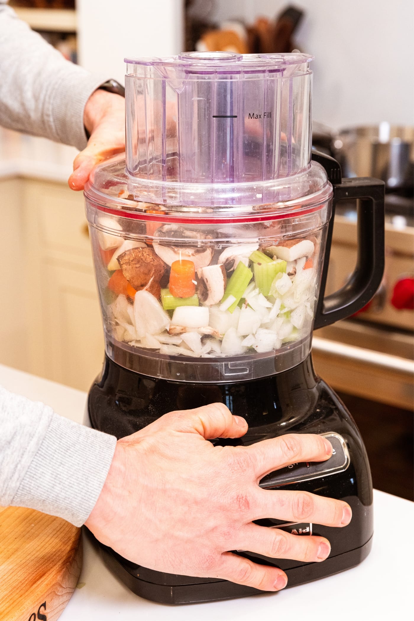 Finer about to push the button on a food processor filled with vegetables including onion, celery, carrots, and mushrooms.