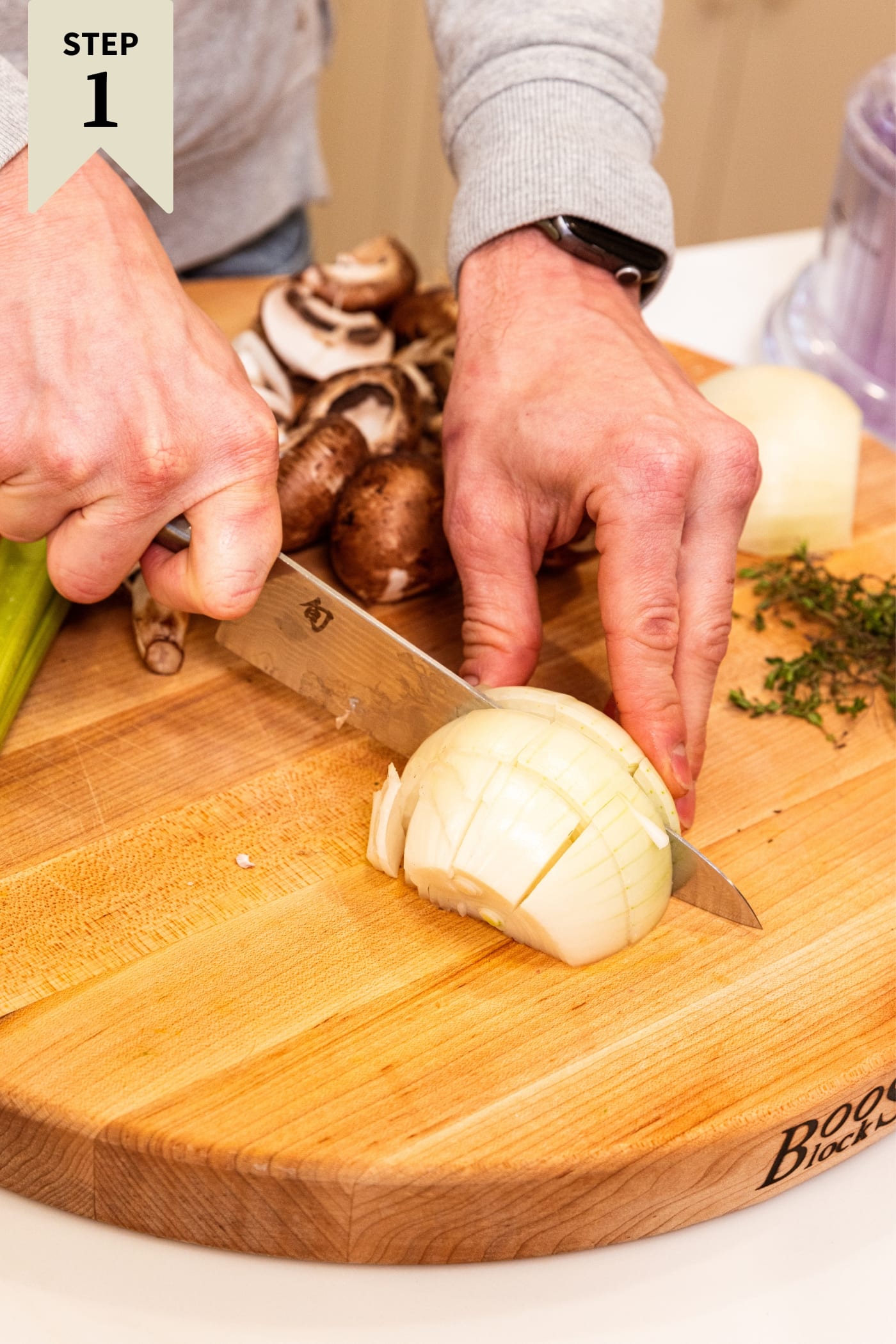 Hand using knife to slice an onion into large chunks on a wooden cutting board with other vegetables in background before being cut.