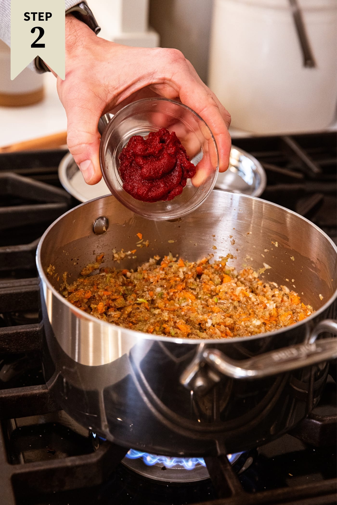 Hand holding small glass bowl about to pour tomato paste into vegetable mixture in a metal stockpot on the heat on stovetop.