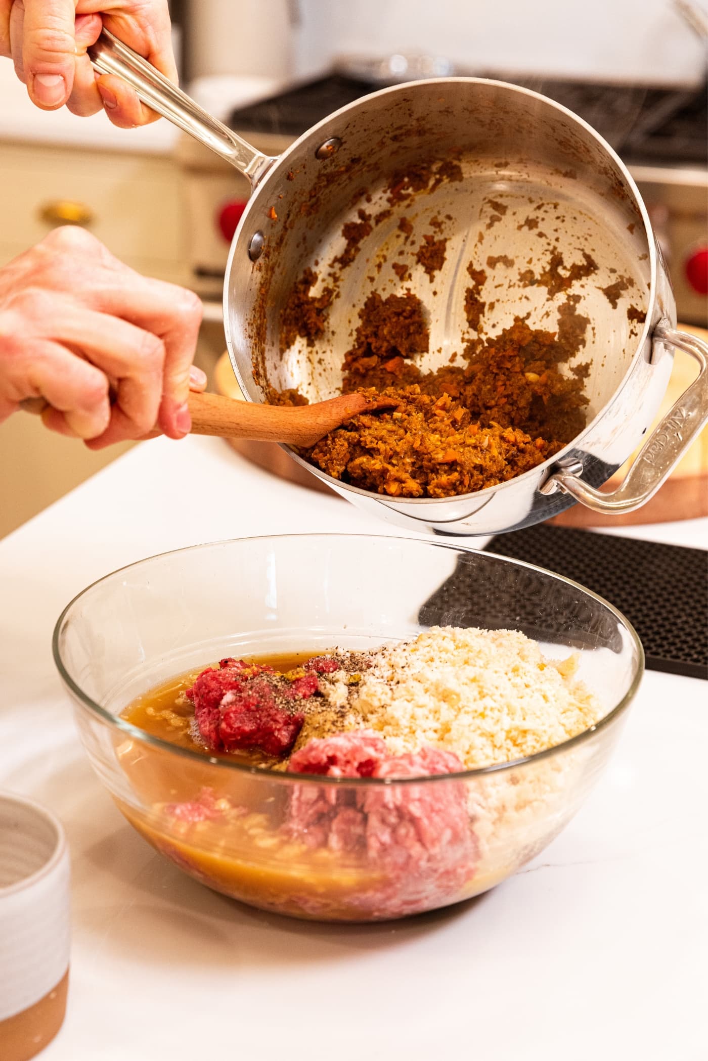 Hand scooping out vegetable mixture from metal stockpot with wooden spoon on top of glass bowl filled with meatloaf ingredients.
