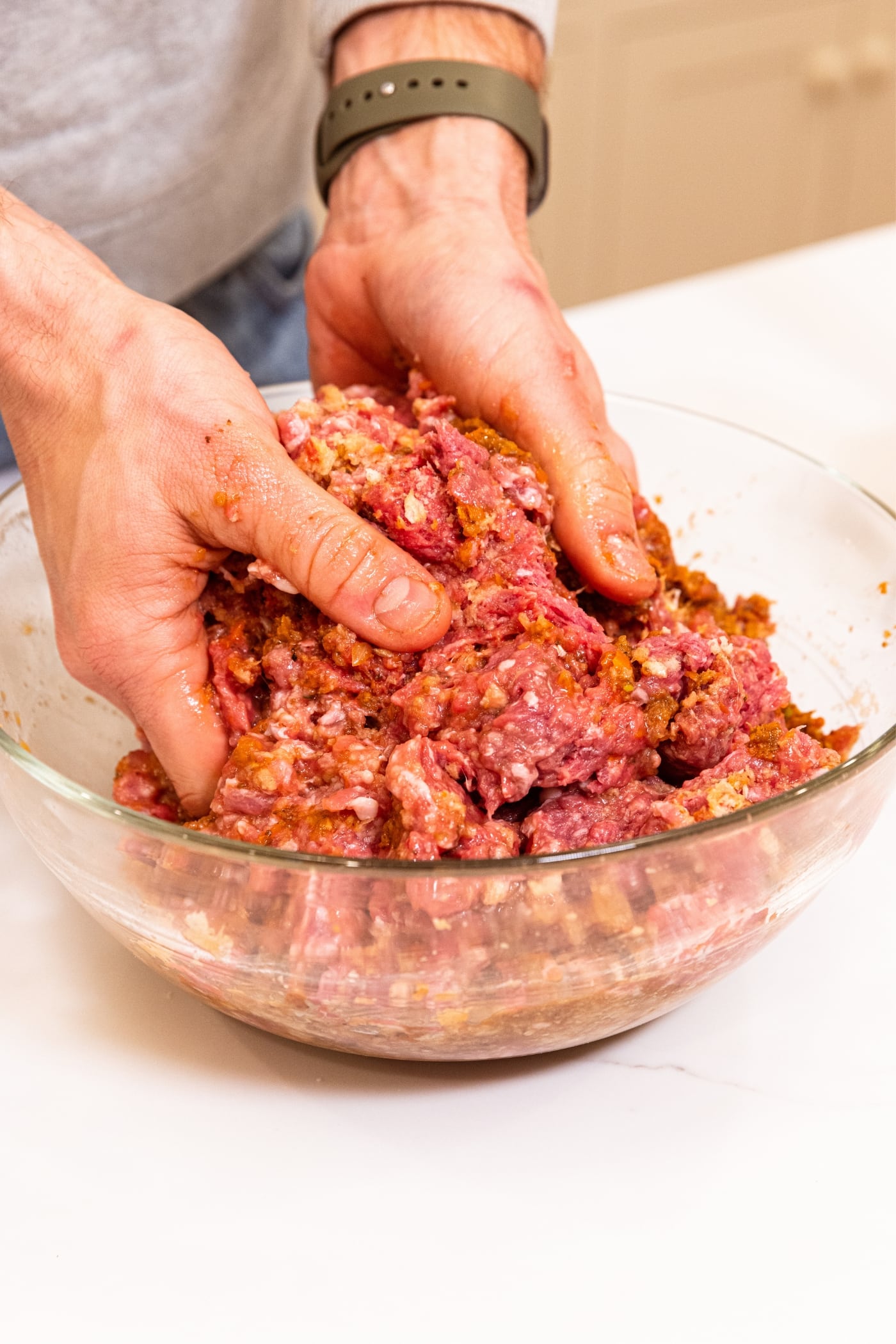 Hands pushing together ingredients needed for meatloaf including beef, pork, and breadcrumbs in glass bowl on white countertop.