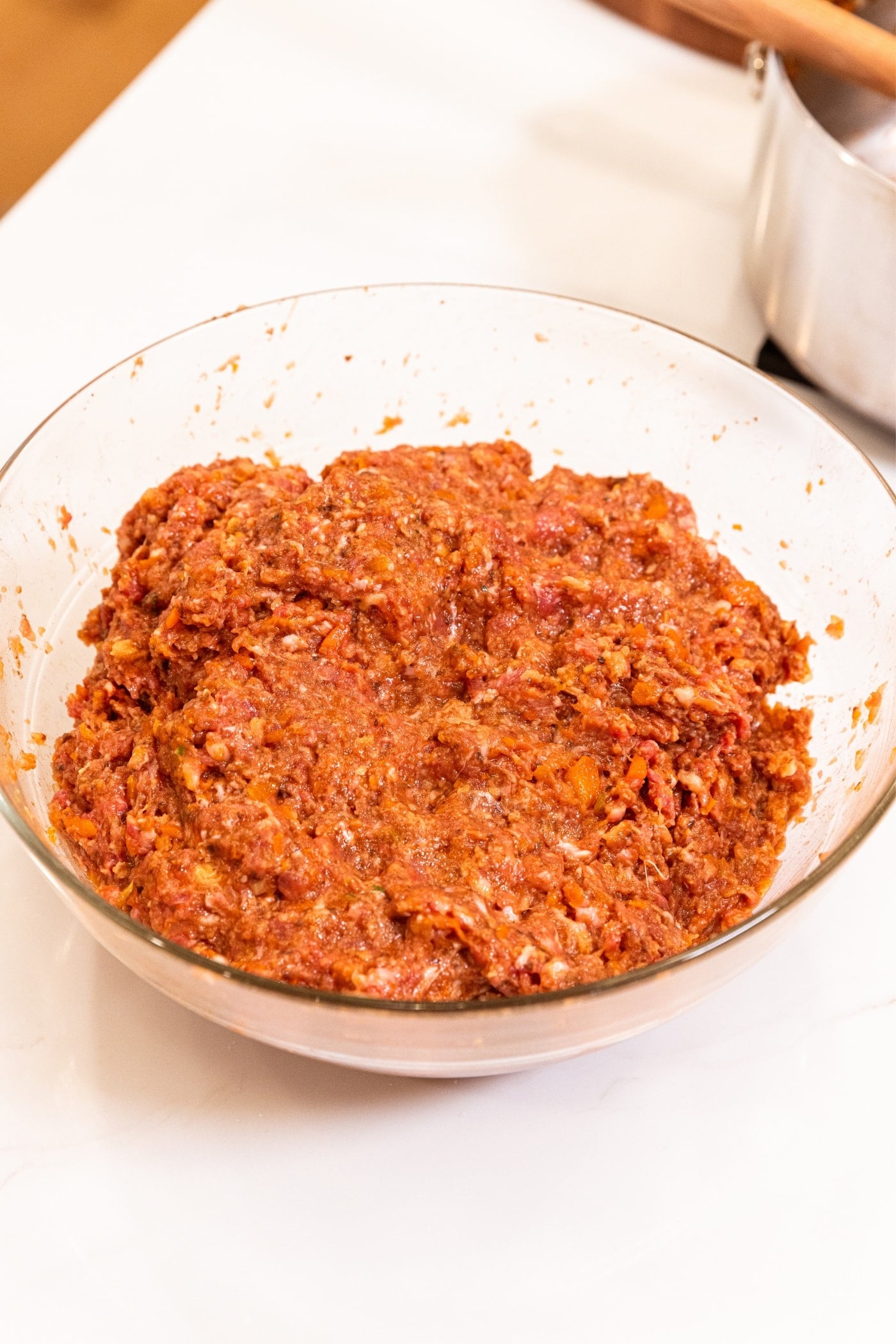 Glass metal bowl with mixed meat mixture for meatloaf sitting on white surface.