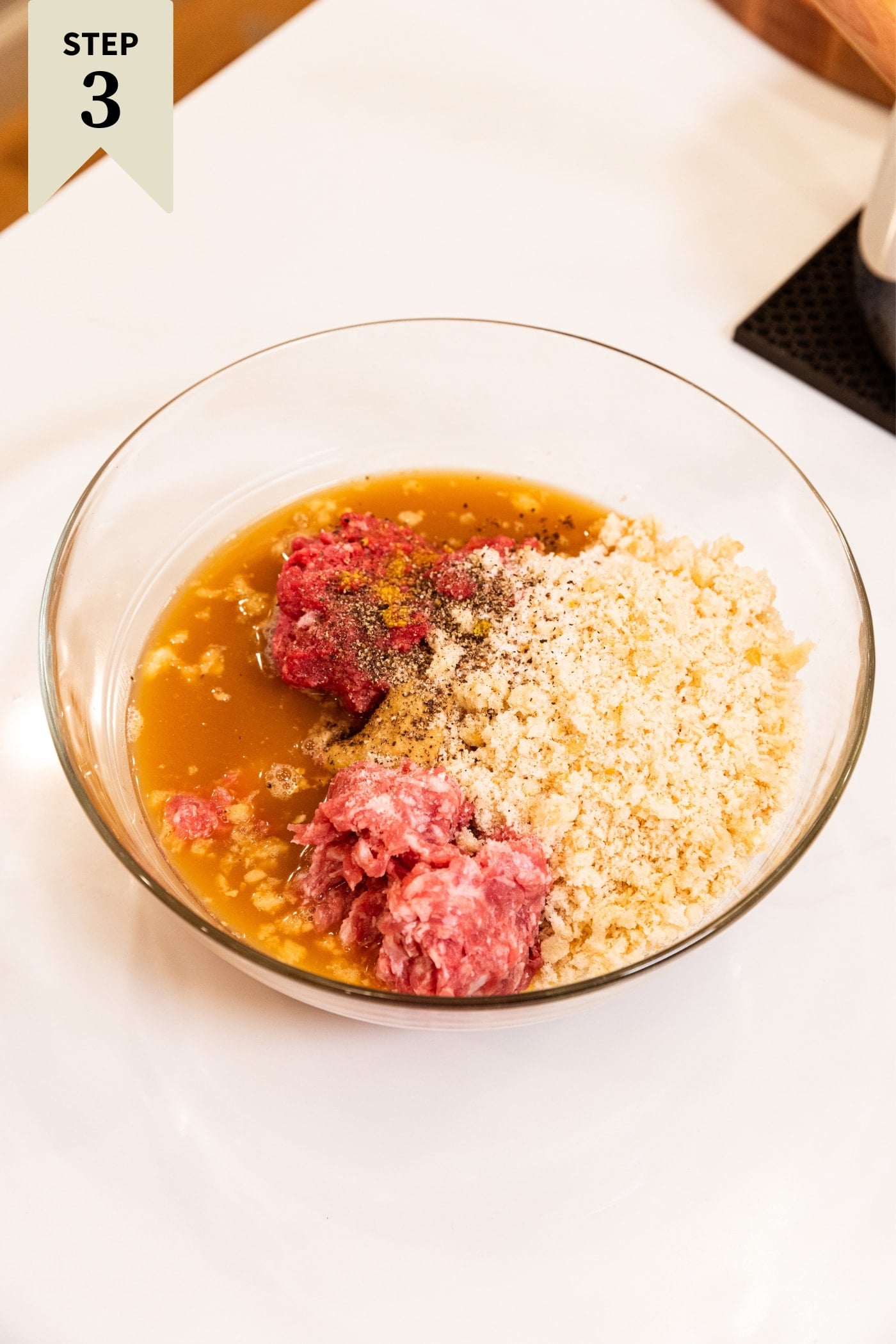Glass bowl filled with all ingredients needed for meatloaf including ground meat, liquids, breadcrumbs, ketchup, and more sitting on white countertop.