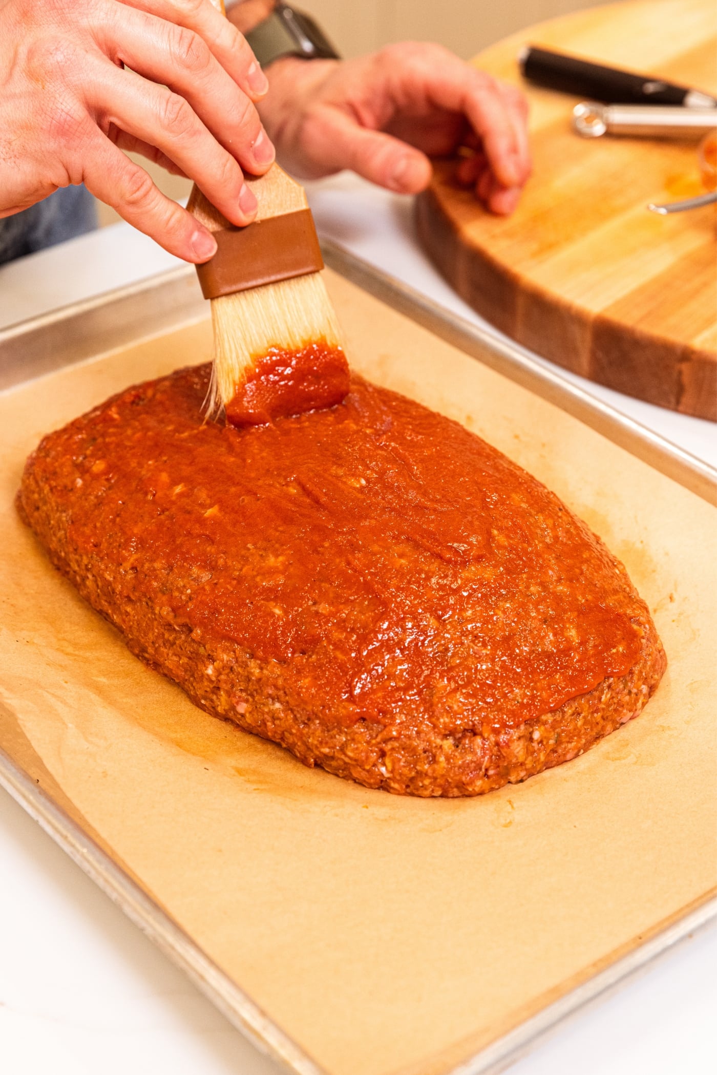 Hand using brush to apply ketchup mixture on the top of meatloaf sitting on parchment-lined baking sheet.