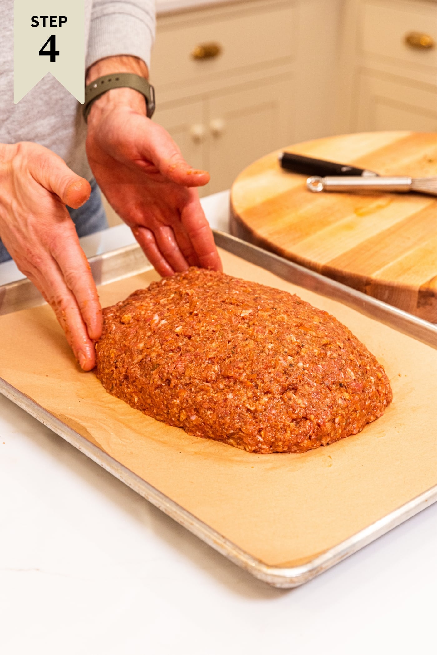 Two hands shaping meatloaf on parchment-lined baking sheet with wood cutting board in background.