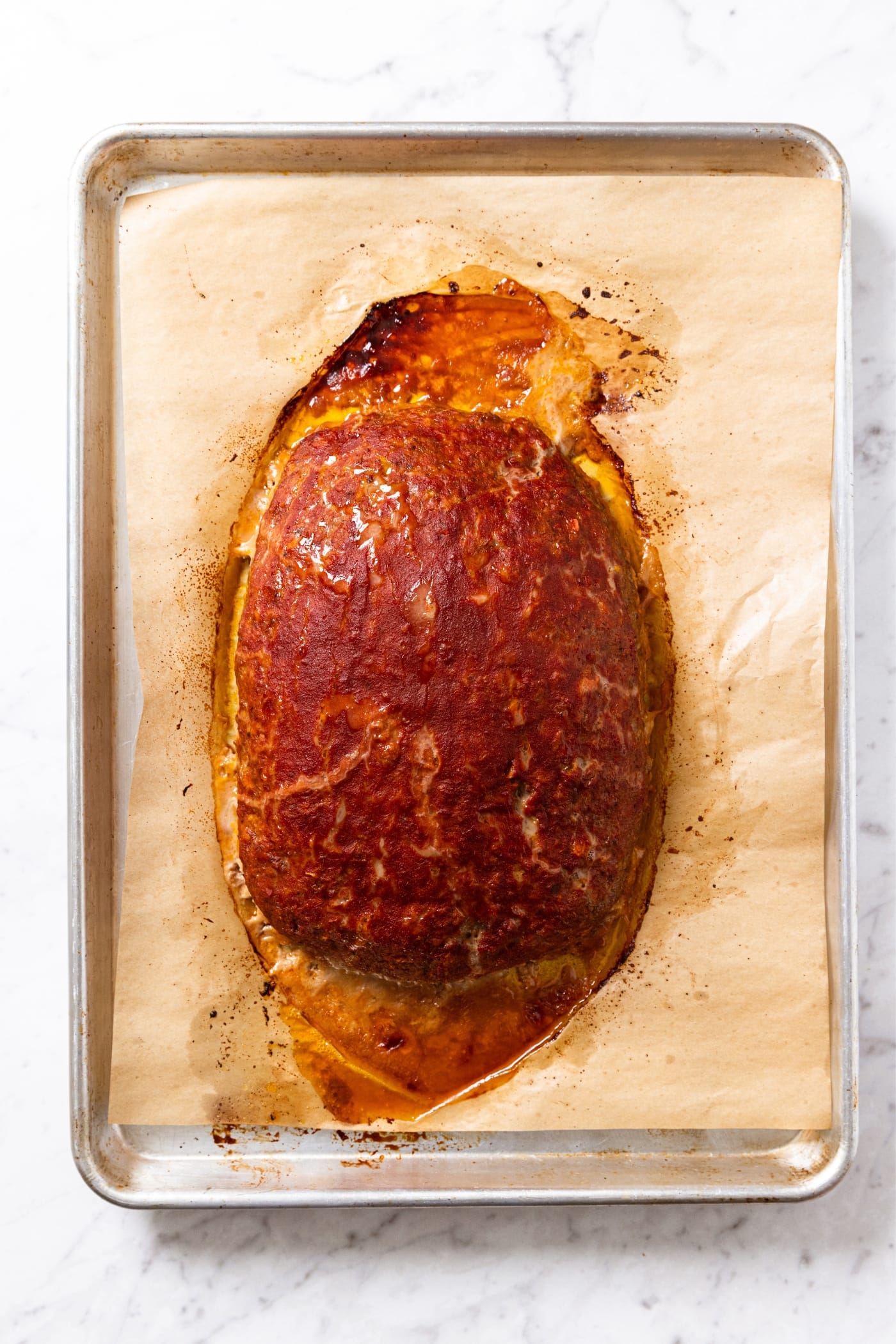 Baked meatloaf sitting on parchment-lined baking sheet with browned ketchup mixture and excess grease from meat seeping out.