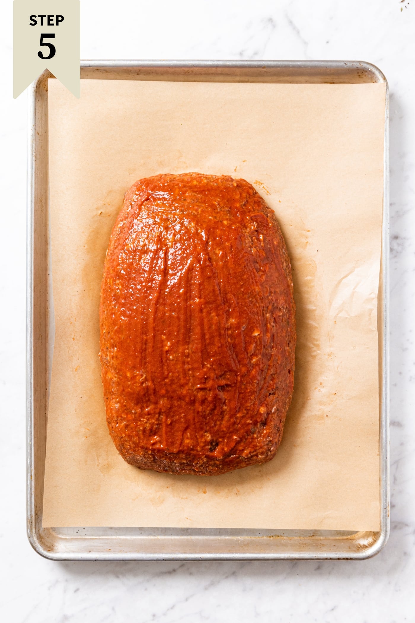 Parchment-lined baking sheet with unbaked meatloaf with ketchup brushed over the top sitting on marble surface.