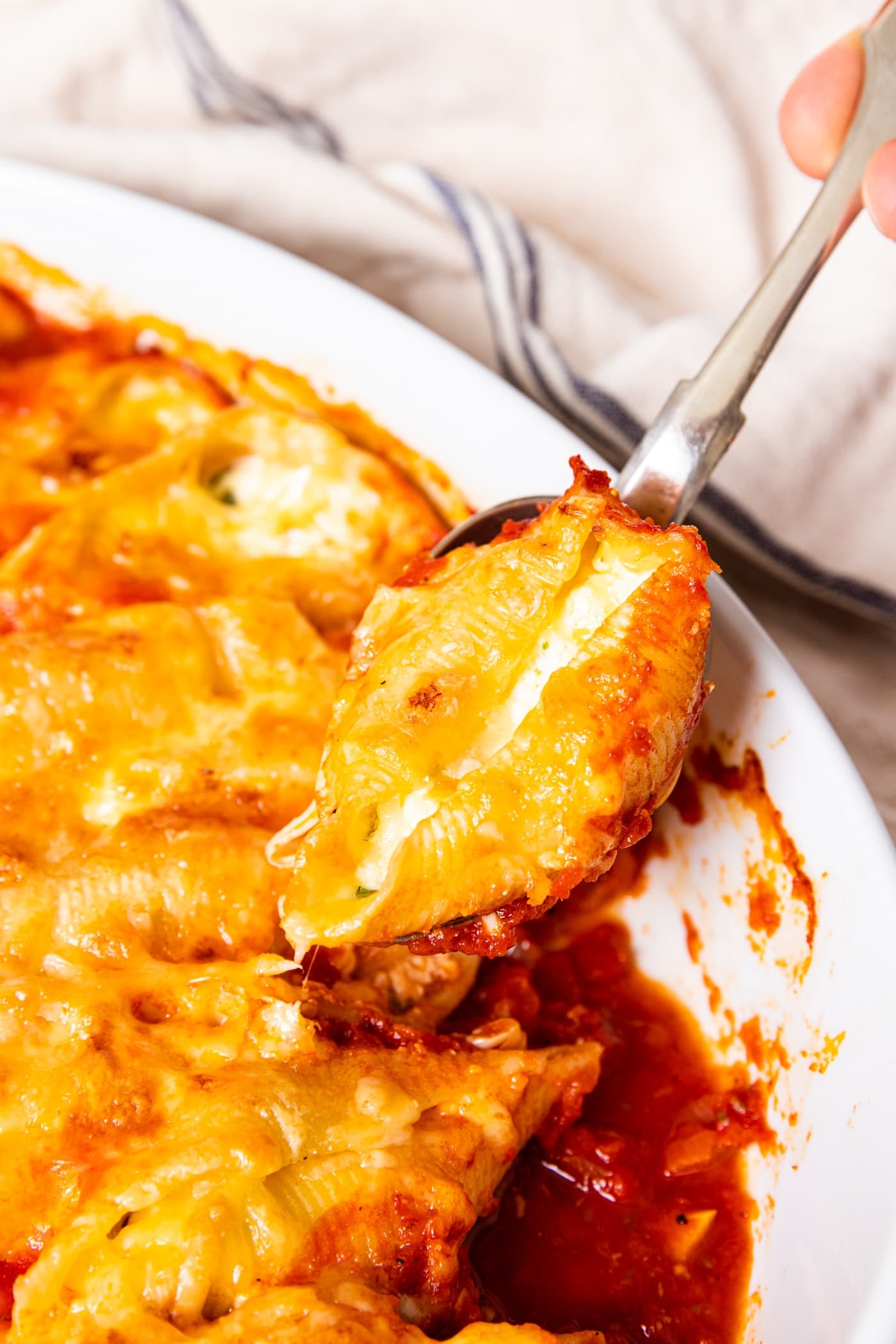White baking dish with stuffed shells in a white baking dish with a spoon holding one shell showing the ricotta mixture inside with tomato sauce underneath.