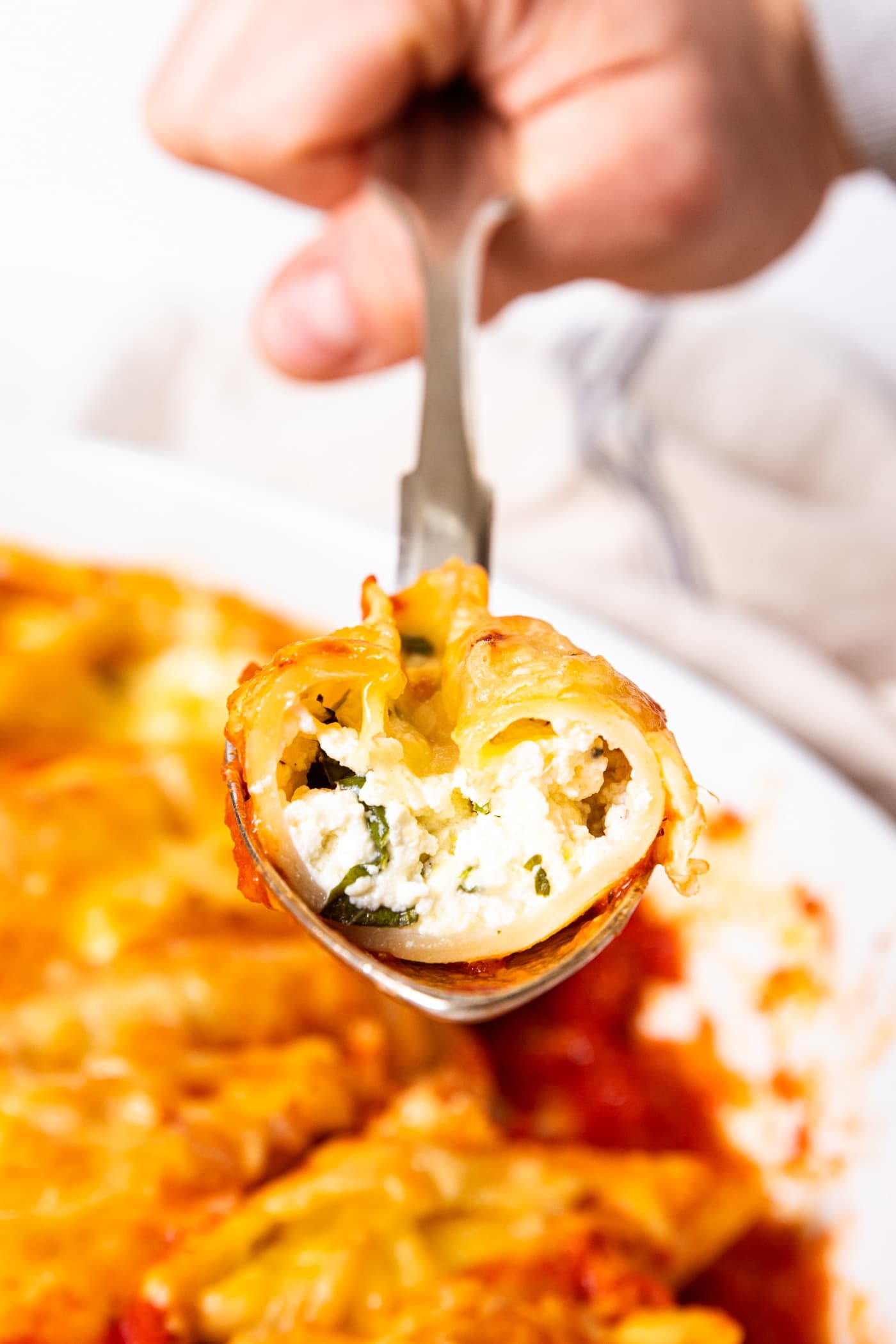 Stuffed shell being held on a silver spoon above the rest of pasta with half cut off showing the interior creamy ricotta mixture inside.