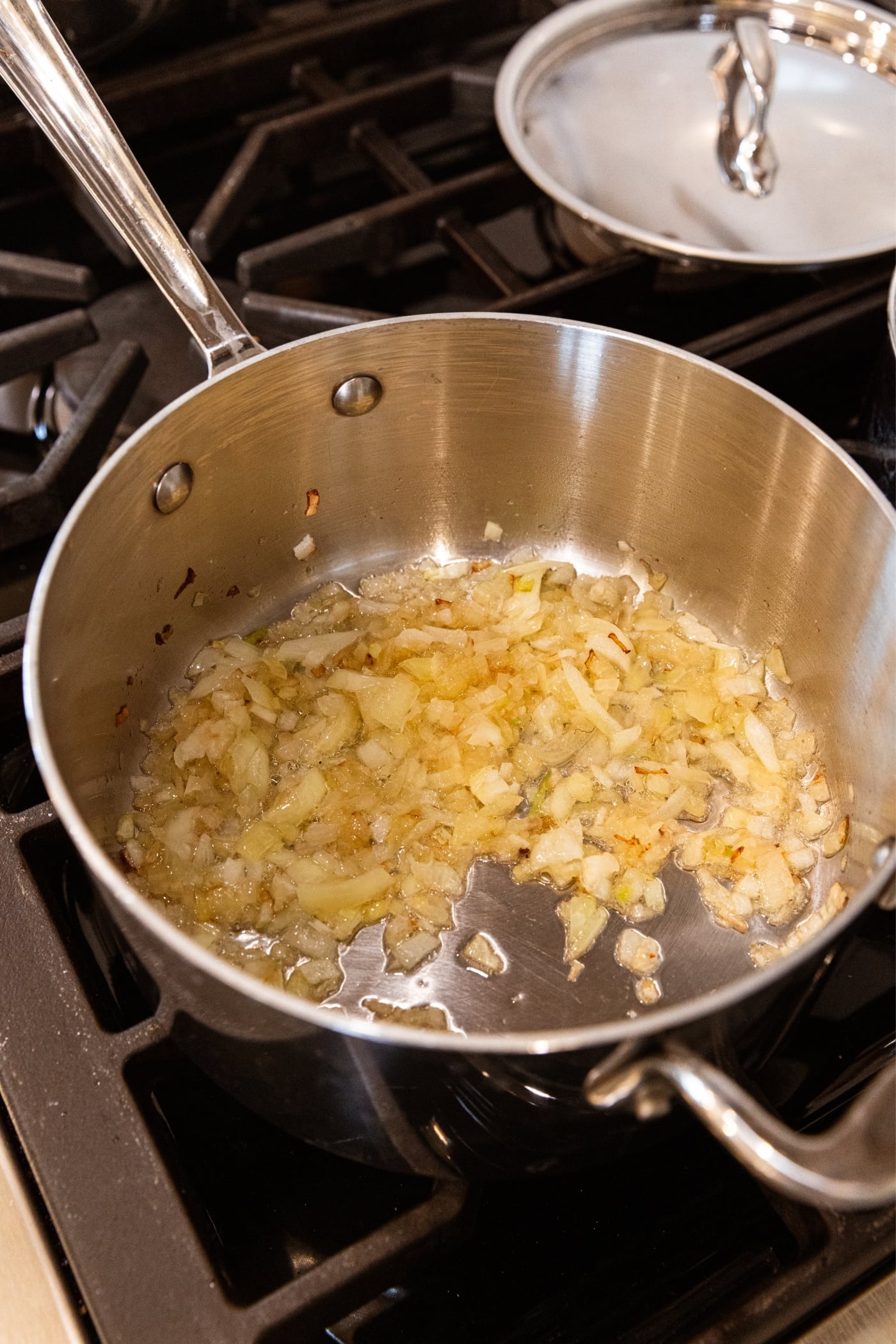 Stainless steel pot sitting on stovetop with browning onion mixture inside with metal lid in background.