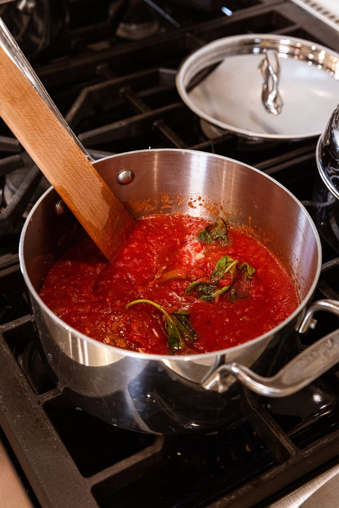 Wooden spatula resting on bottom of inside of metal pot filled with tomato sauce with a sprig of basil showing through the sauce being heated on the stovetop.