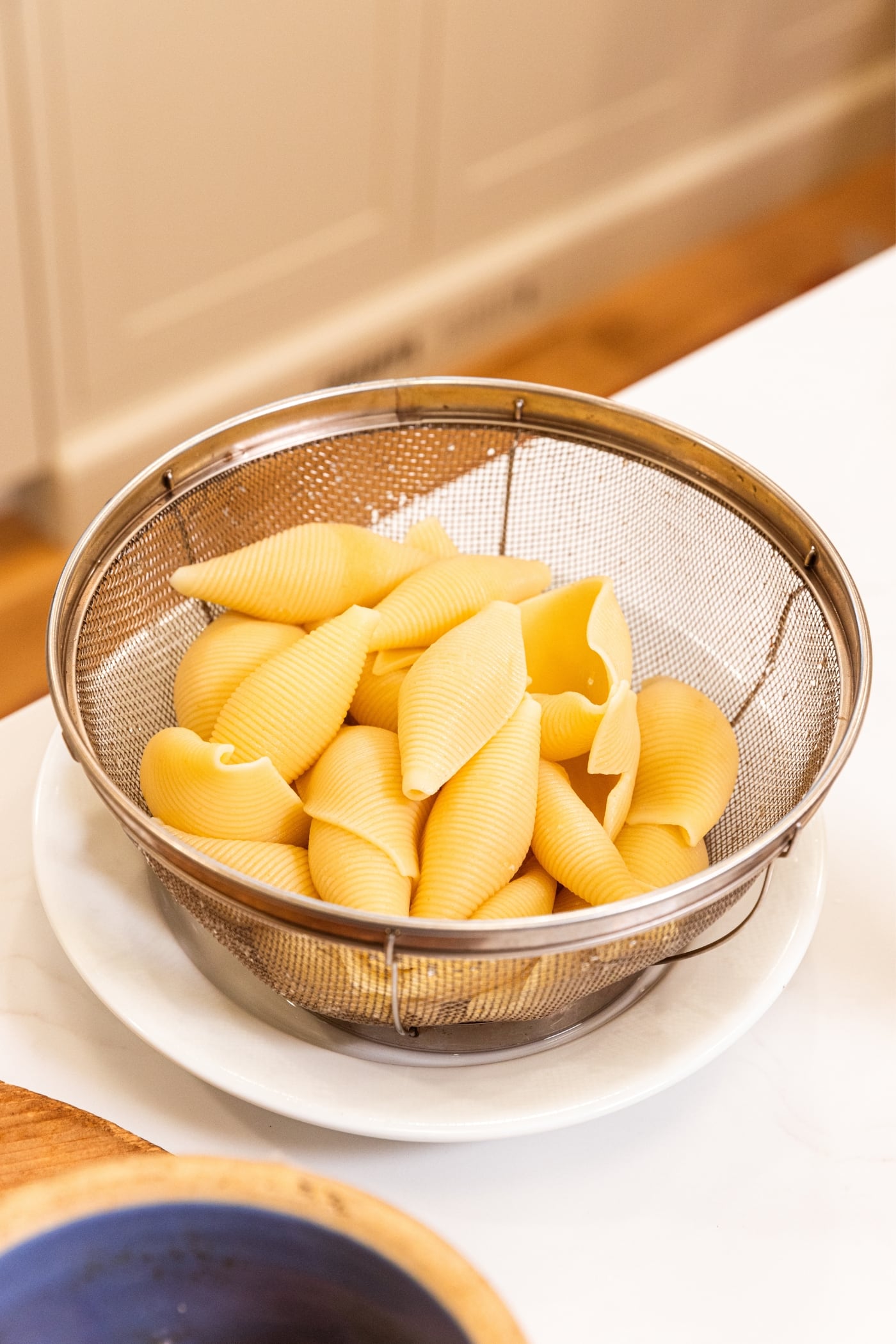 Metal colander sitting on white plate on countertop after being drained out of stockpot and then rinsed waiting to be filled as stuffed shells.