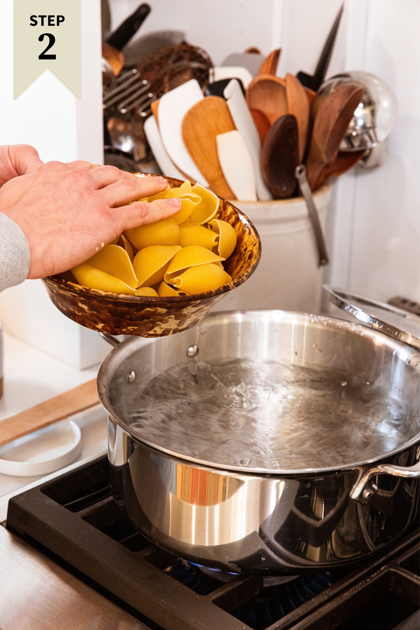 Hand holding brown bowl filled with pasta shells about to pour them into a large stockpot filled with boiling water on stovetop.