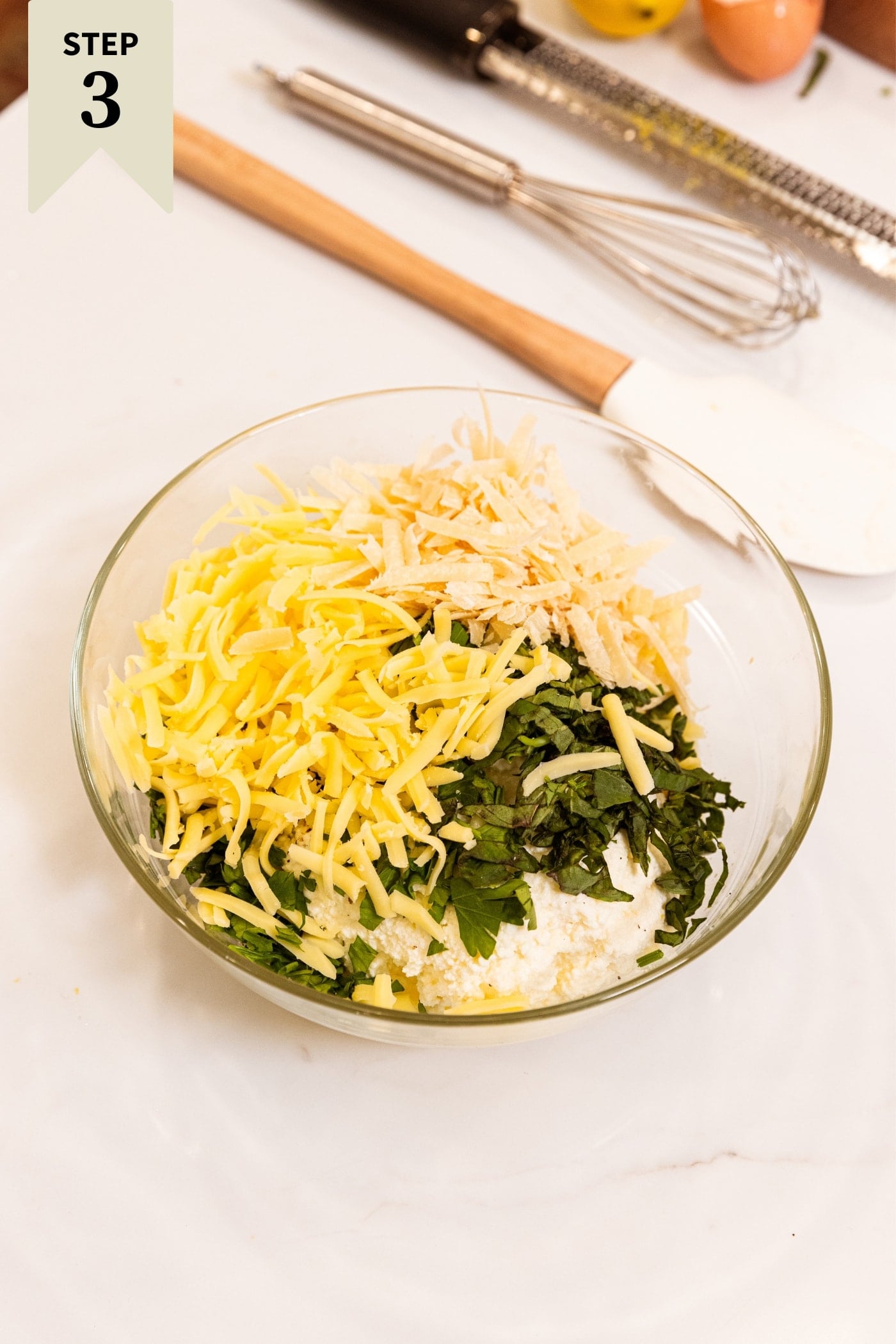 Glass bowl filled with ingredients needed to make ricotta cheese filling for stuffed shells sitting on white surface with mixing utensils in background.