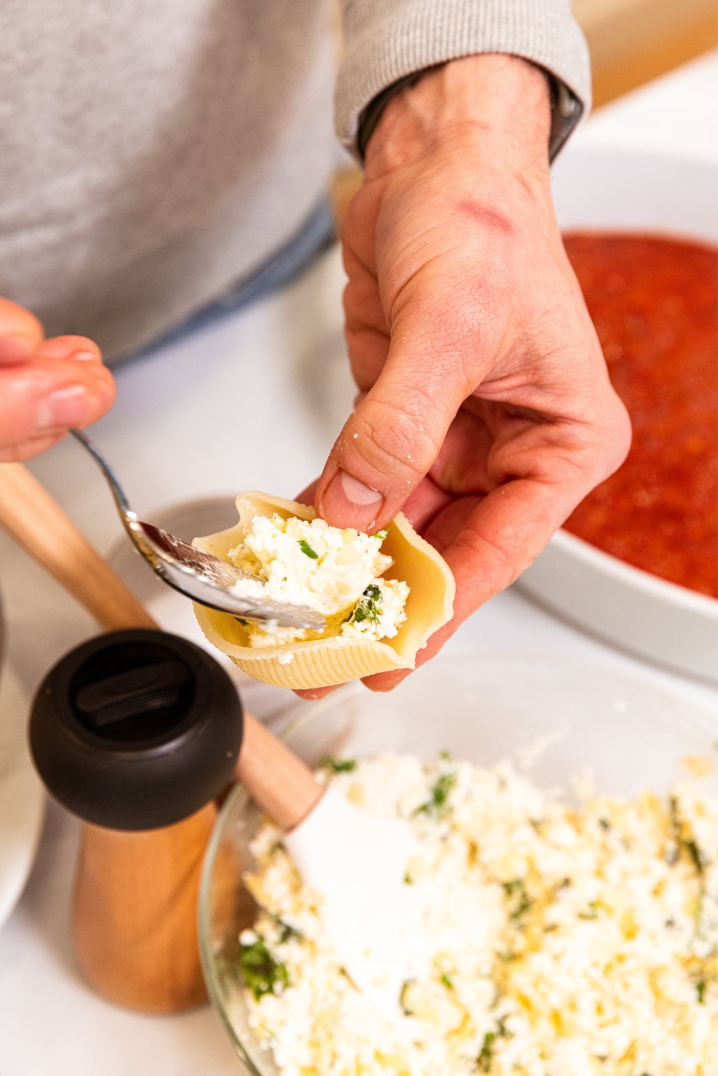 Hand holding pasta shell with other hand holding spoon putting ricotta mixture inside shell with rest of ricotta mixture in glass bowl below.