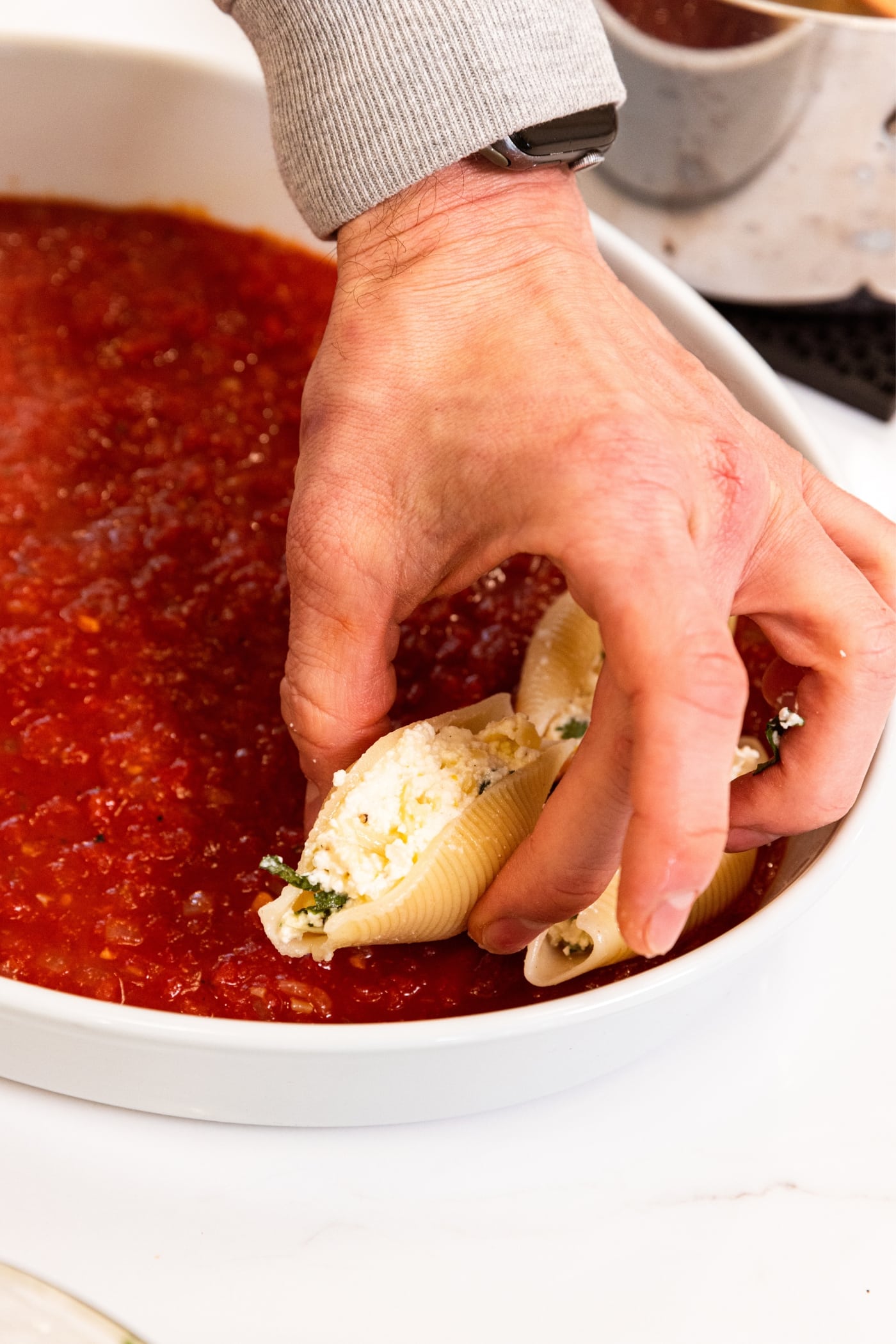 Hand placing stuffed shell into white baking dish with tomato sauce all over the bottom of the pan.