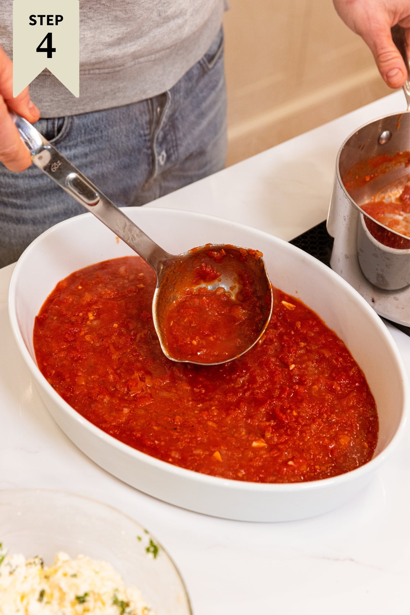 White oval baking dish filled with tomato sauce with hand holding ladle spooning more into bottom of pan.