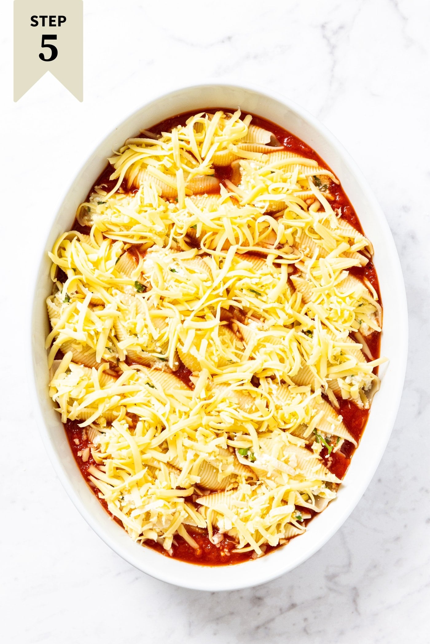 White oval baking dish sitting on marble surface with stuffed shells sitting on tomato sauce with cheese sprinkled over the top right before going into the oven to bake.
