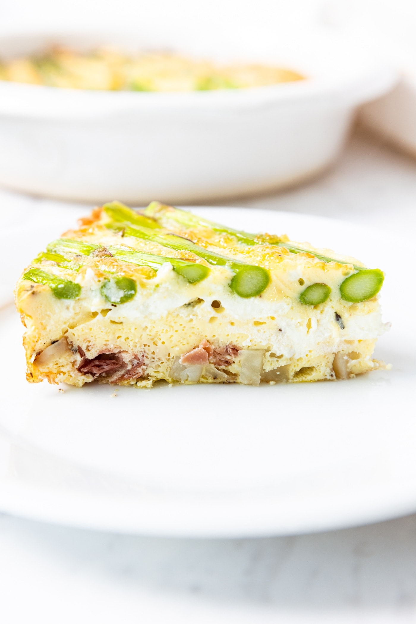 White plate with slice of asparagus and roasted potato frittata with goat cheese and slices of asparagus on top with pie dish in background.
