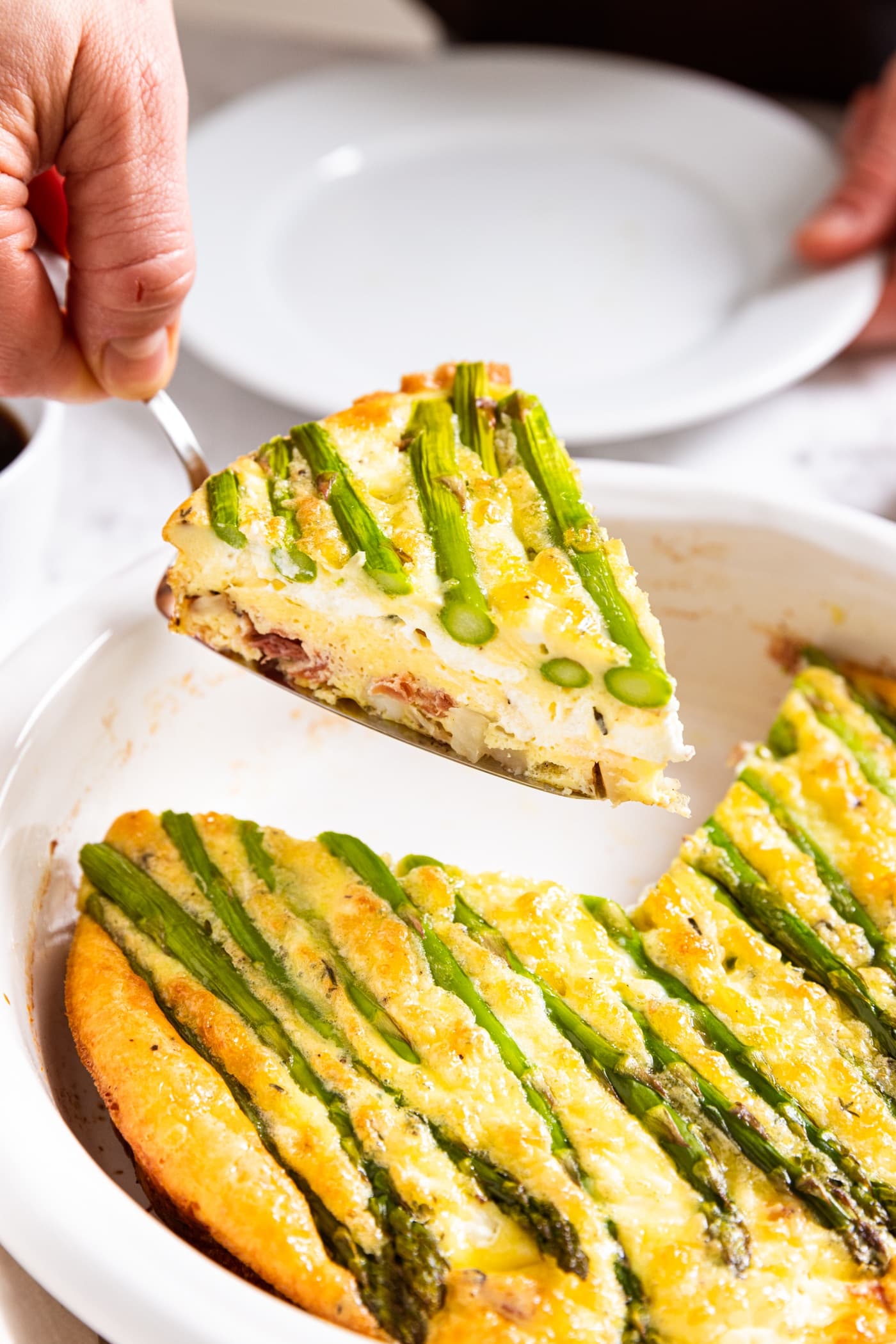Hand using scoop to hold up a slice of asparagus and potato frittata from white pie dish filled with the rest of the frittata.