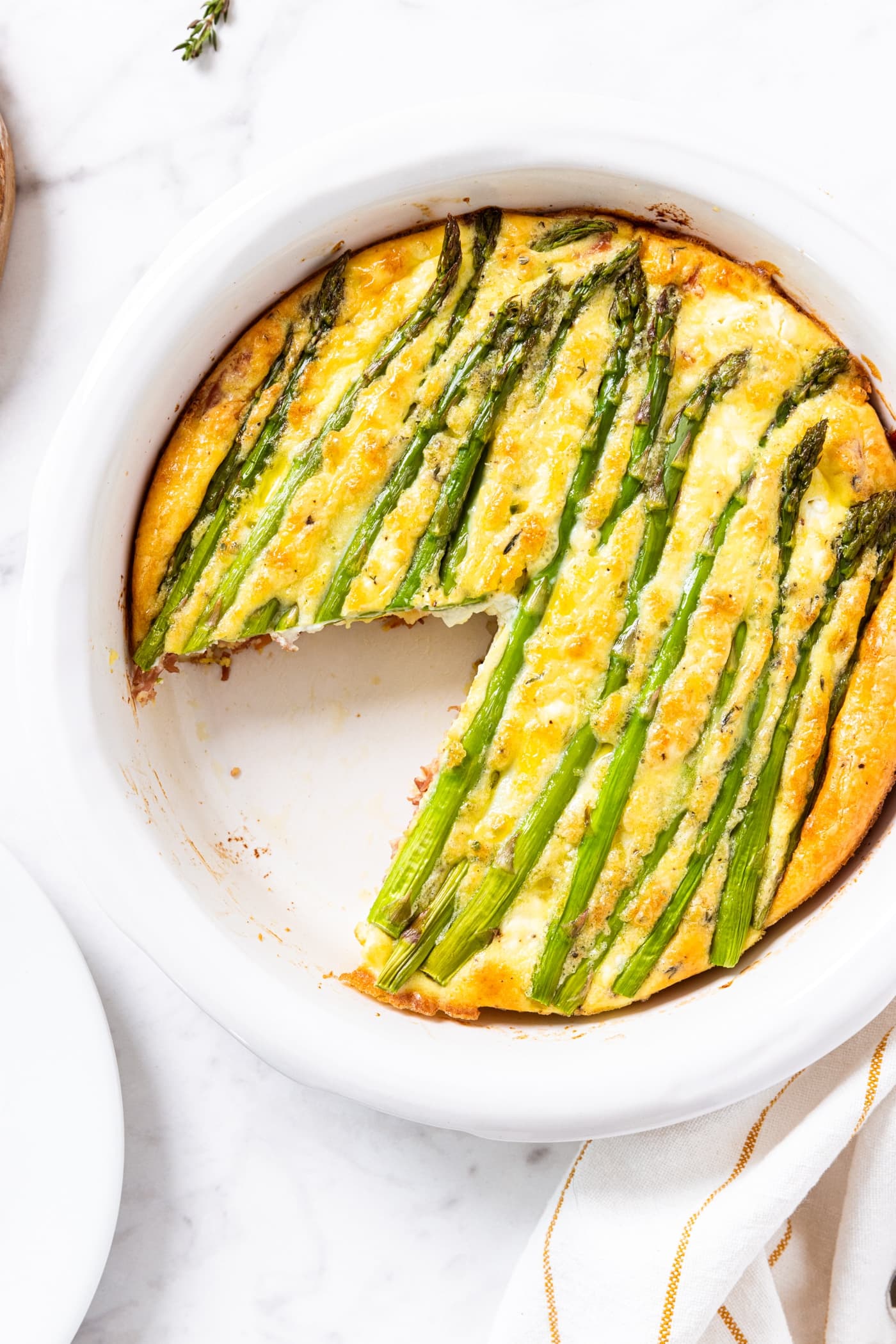 Round white pie dish filled with baked asparagus and roasted potato frittata sitting on white marble surface with one slice removed with stalks of asparagus baked on top.