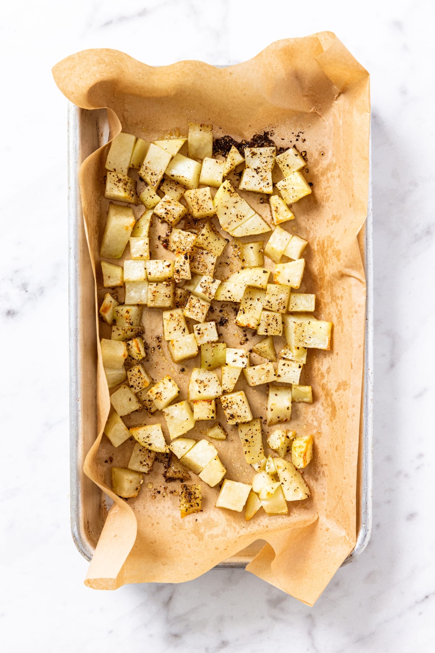 White marble surface with small metal baking sheet with piece of parchment paper with roasted chunks of potatoes right after coming out of the oven.