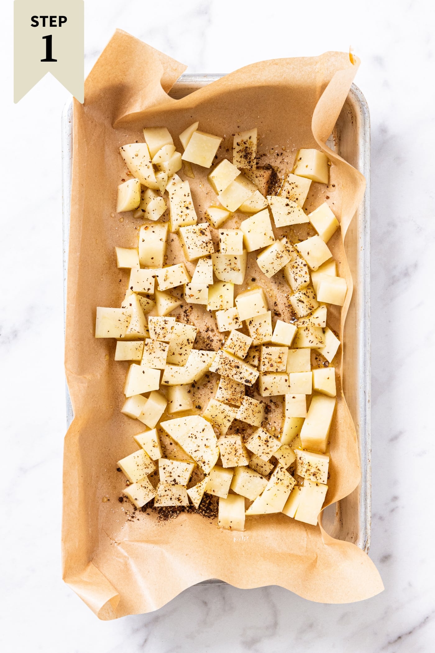 White marble surface with small metal baking sheet with brown-colored parchment paper with unbaked diced potato sprinkled with black pepper and salt right before going into the oven.