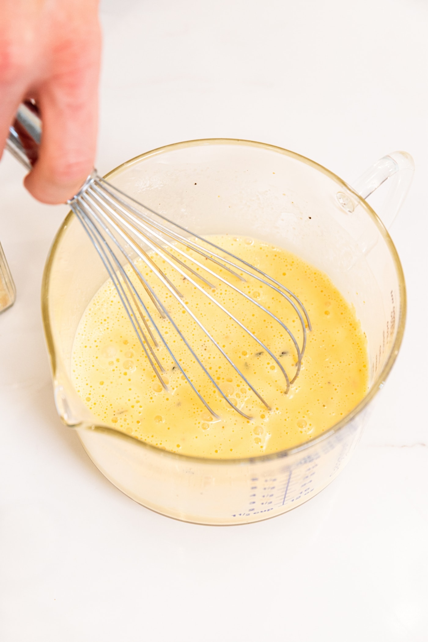 Hand holding whisk mixing together egg and cream mixture in glass measuring cup on white surface.