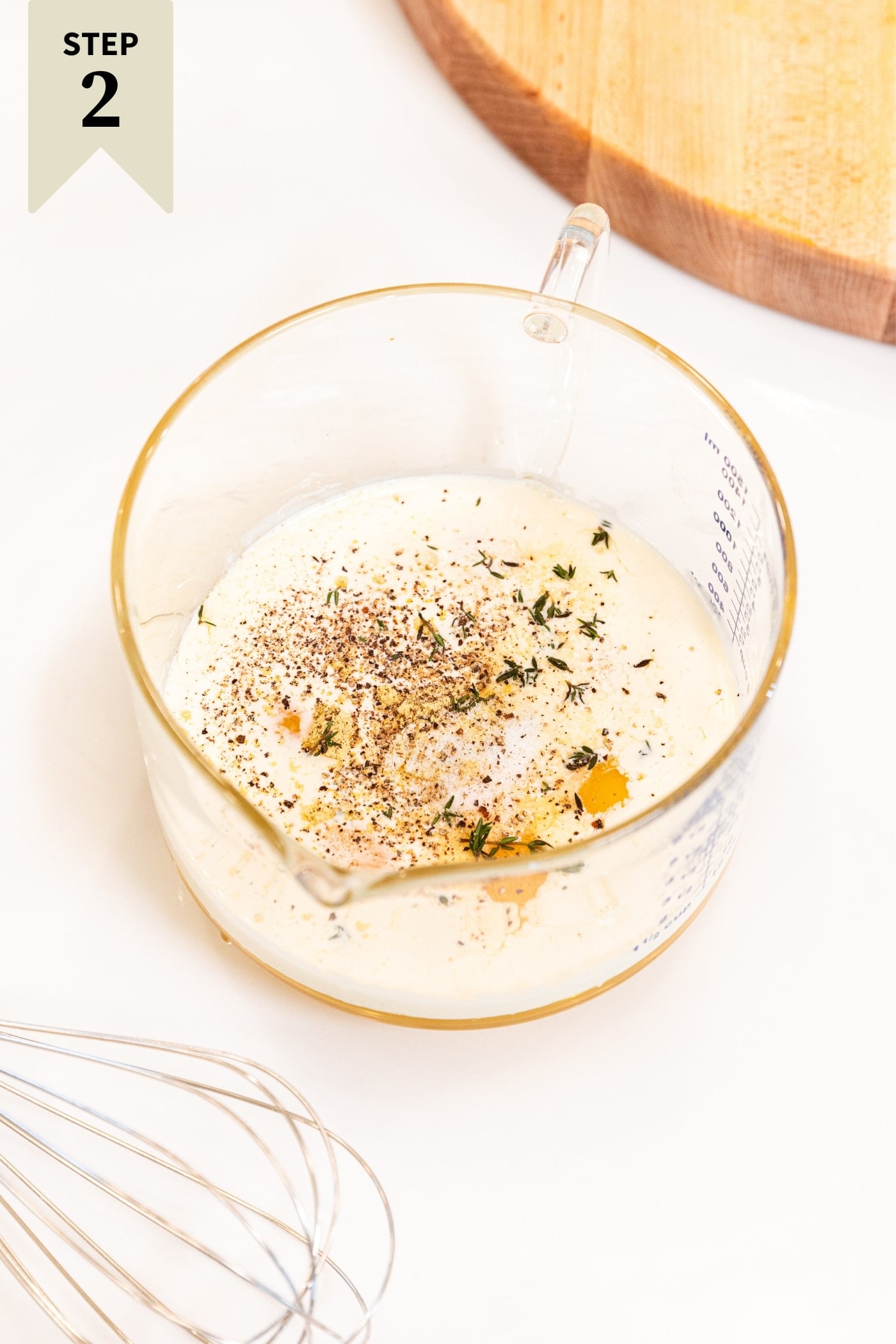 White surface with glass measuring cup filled with eggs, cream, black pepper, and thyme before being mixed together with a whisk.