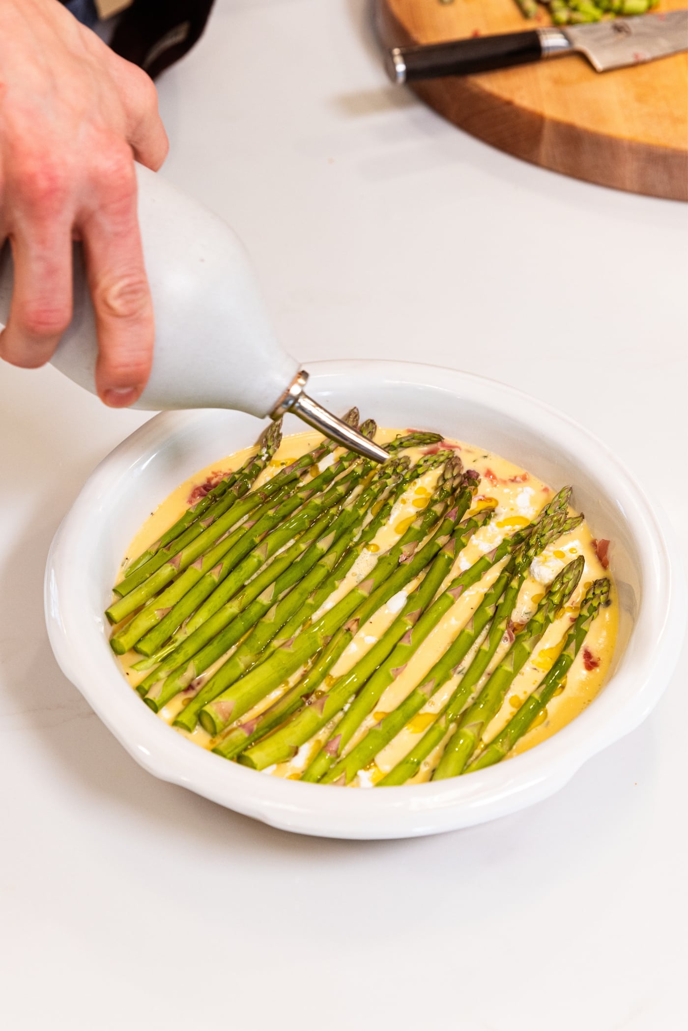 Hand drizzling olive oil from a bottle on top of a white pie dish filled with asparagus on top of an egg mixture on white surface.