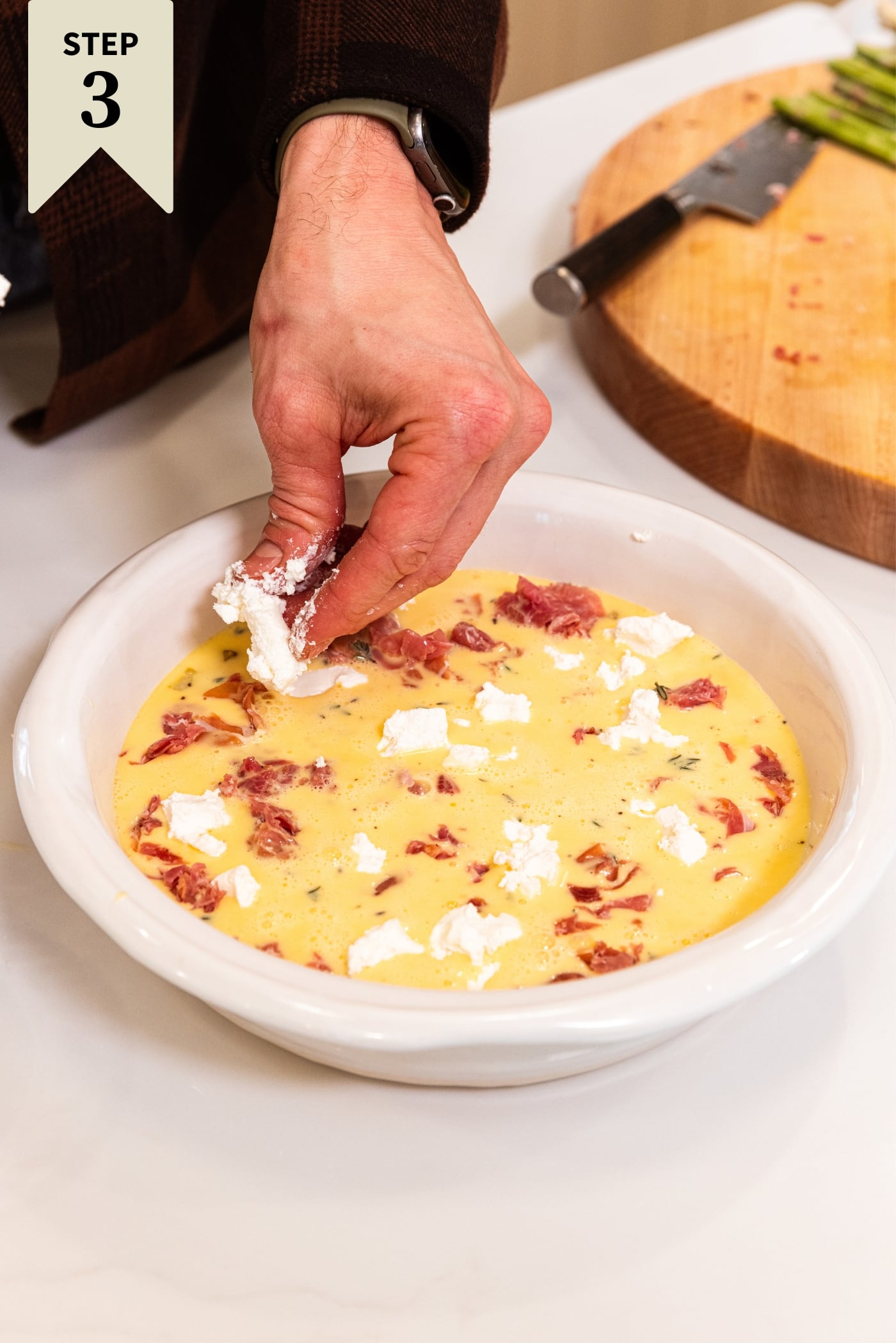 Hand crumbling white goat cheese on the top of an egg mixture in a round white pie dish sitting on white surface.