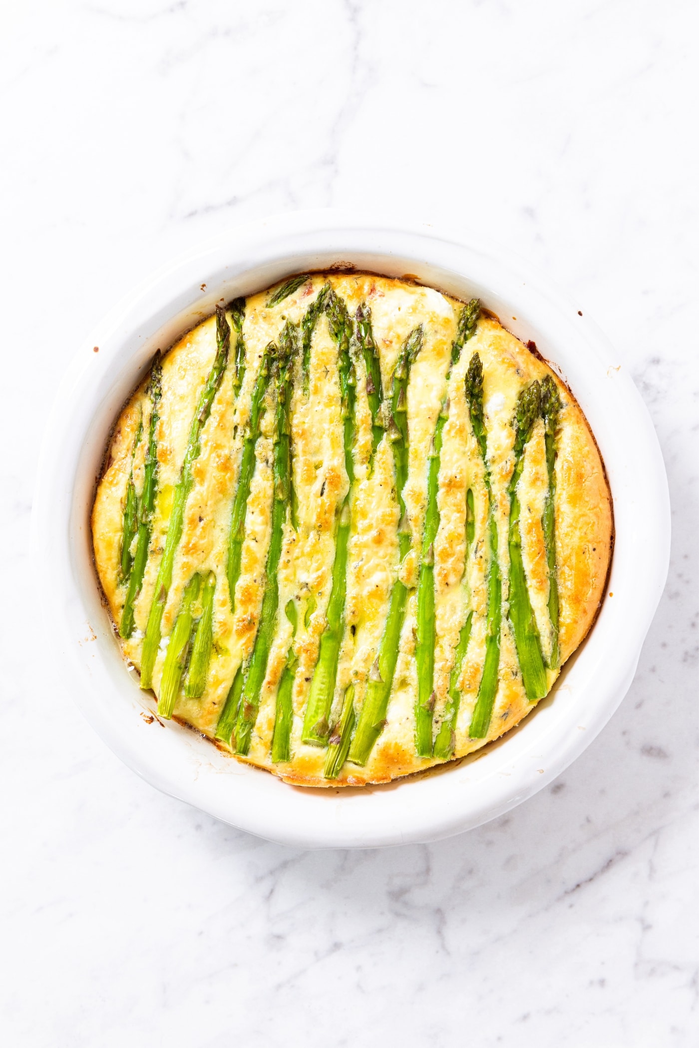 Baked frittata topped with stalks of asparagus right after coming out of the oven sitting on marble surface.
