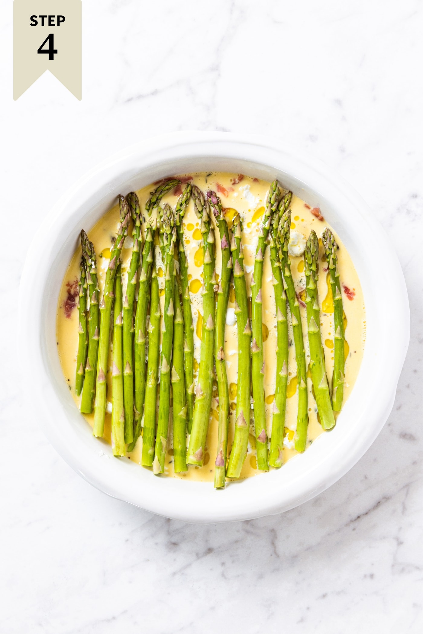 White marble surface with unbaked frittata topped with stalks of asparagus all in white pie dish right before going into the oven to bake,.