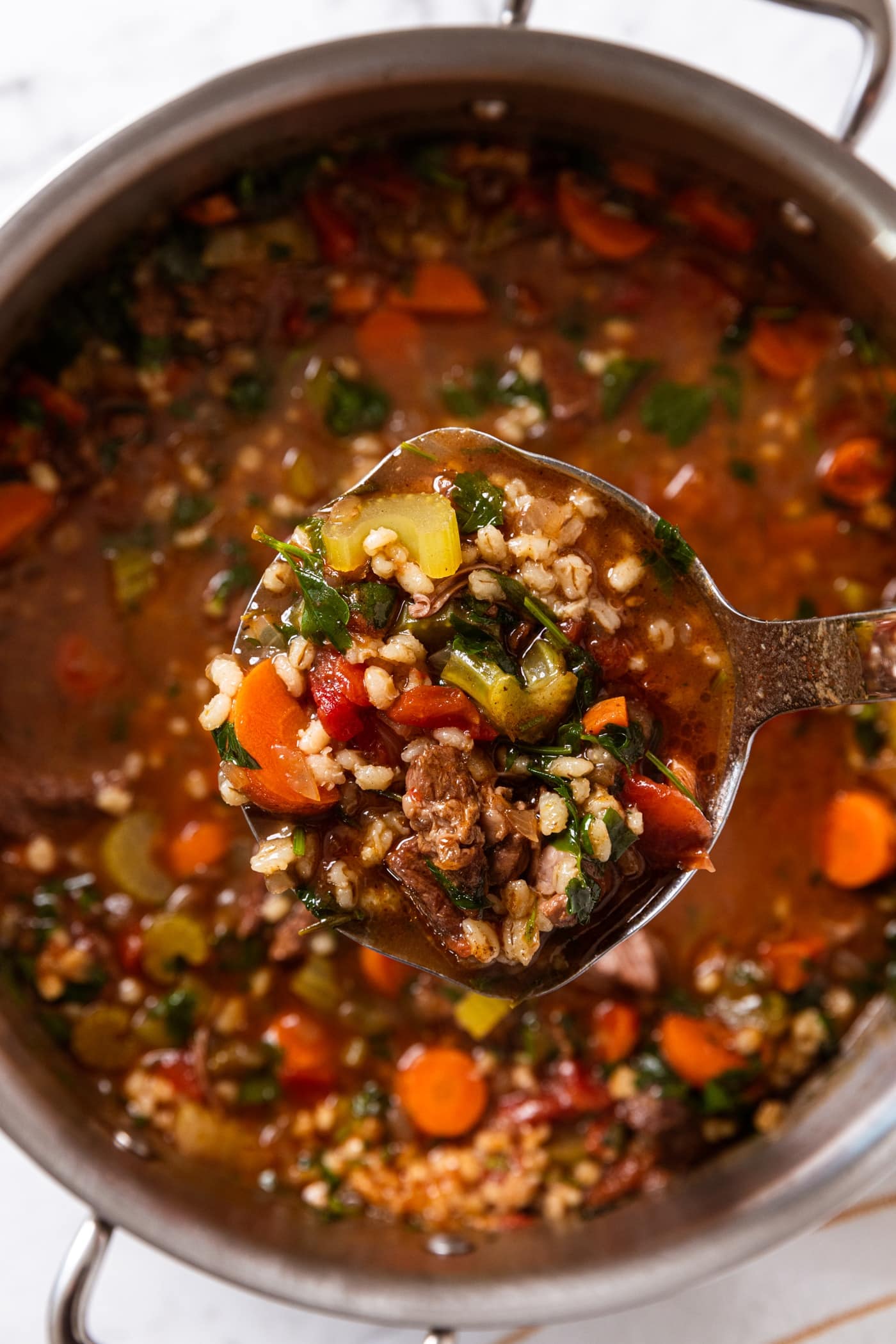 White marble surface with metal stockpot filled with brown-colored beef and barley soup with ladle holding serving of soup showing barley.