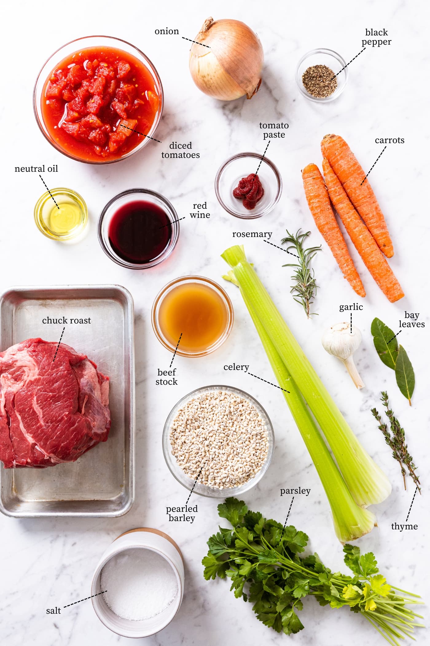 Marble surface filled with all ingredients needed to prepare beef and barley soup including raw chuck roast, celery, carrots, herbs, spices, red wine, barley, onion, and more.