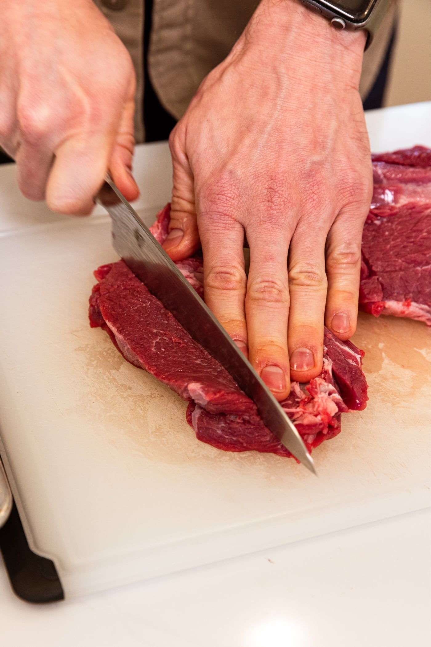 Hand pushing down on one side of strip of raw chuck roast cutting off another strip of meat.