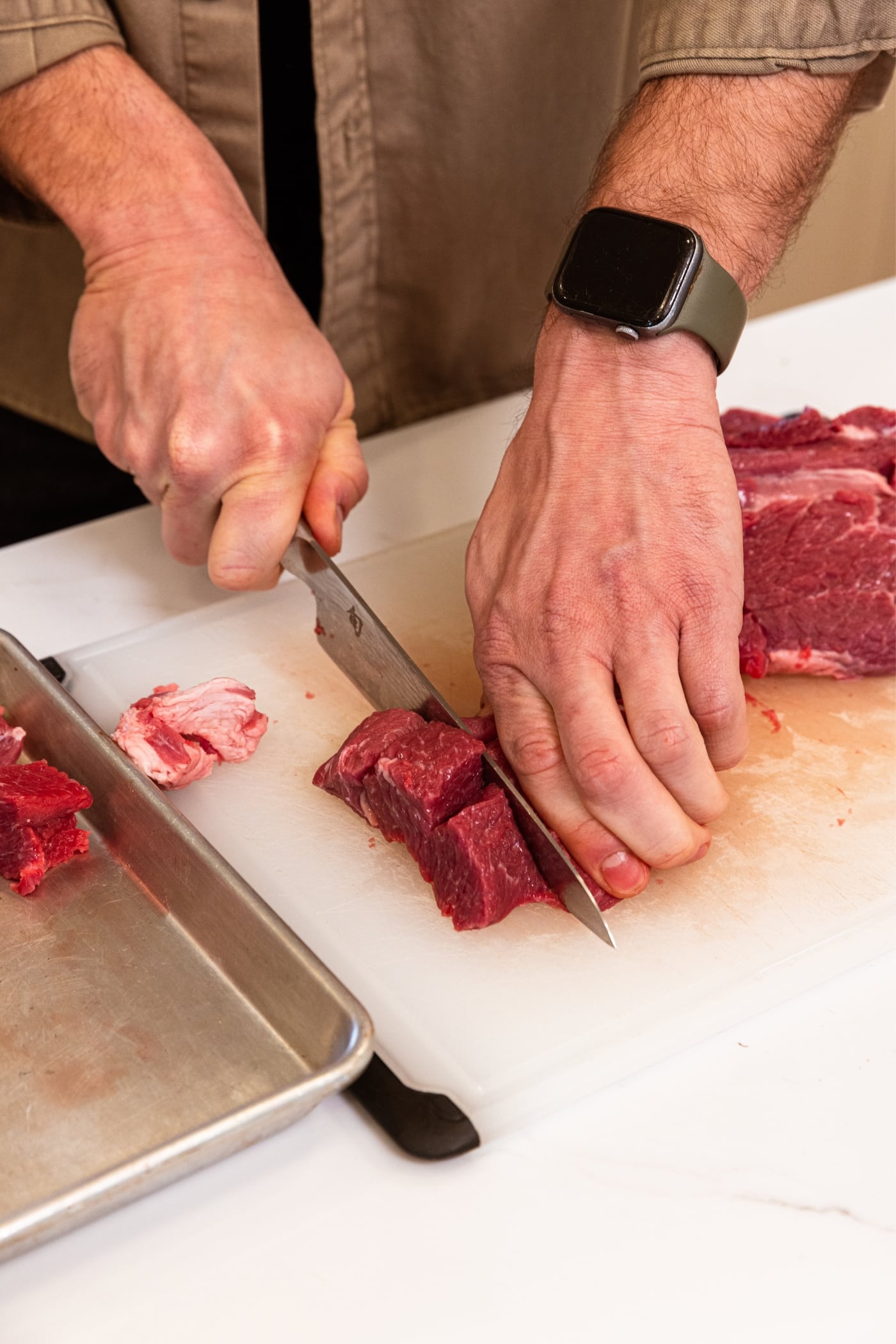 Hand using knife to slice strip of raw chuck roast into small 1-inch cubes on white cutting board.