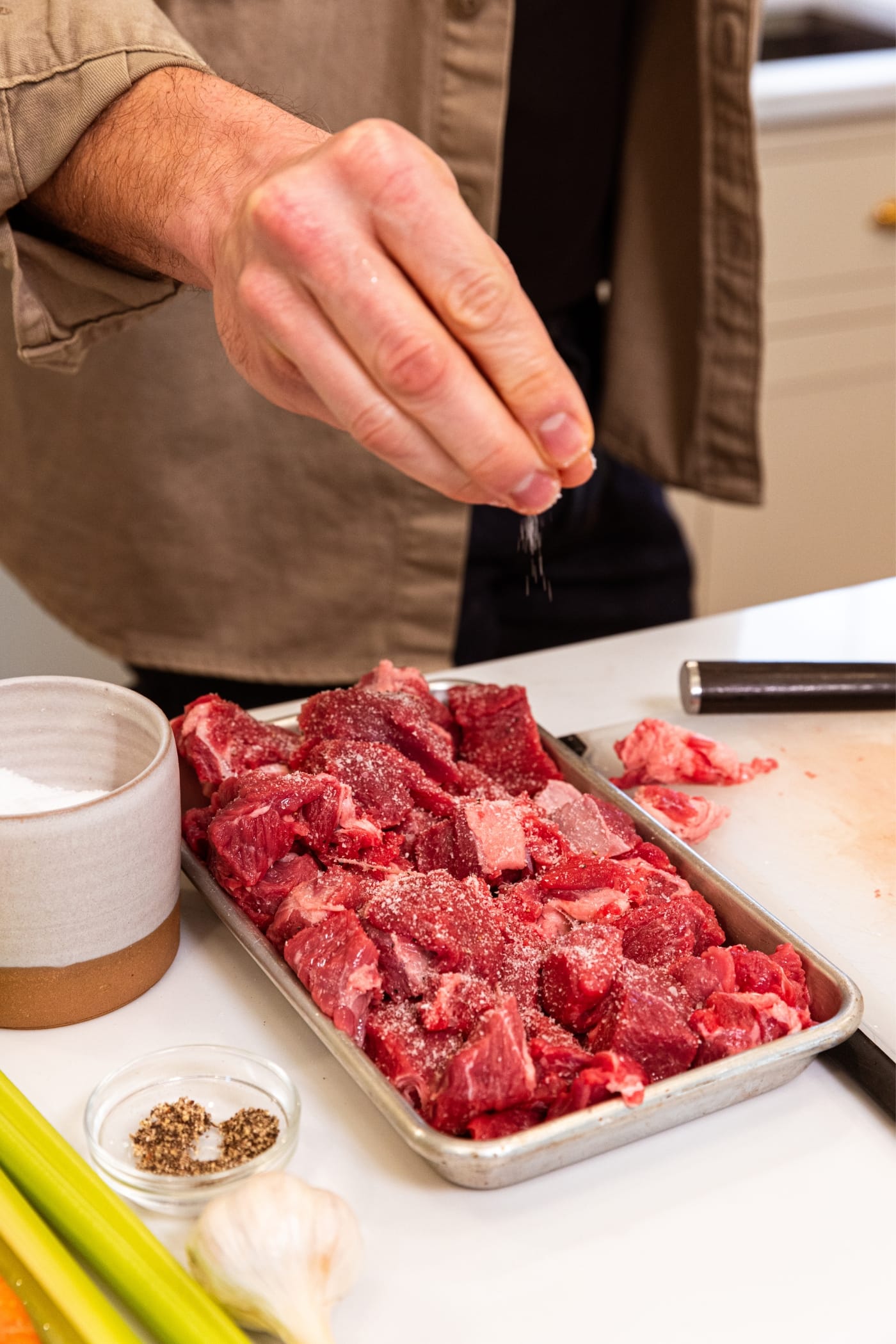 Hand sprinkling salt on top of cubes of raw chuck roast sitting on small metal sheet pan in kitchen.
