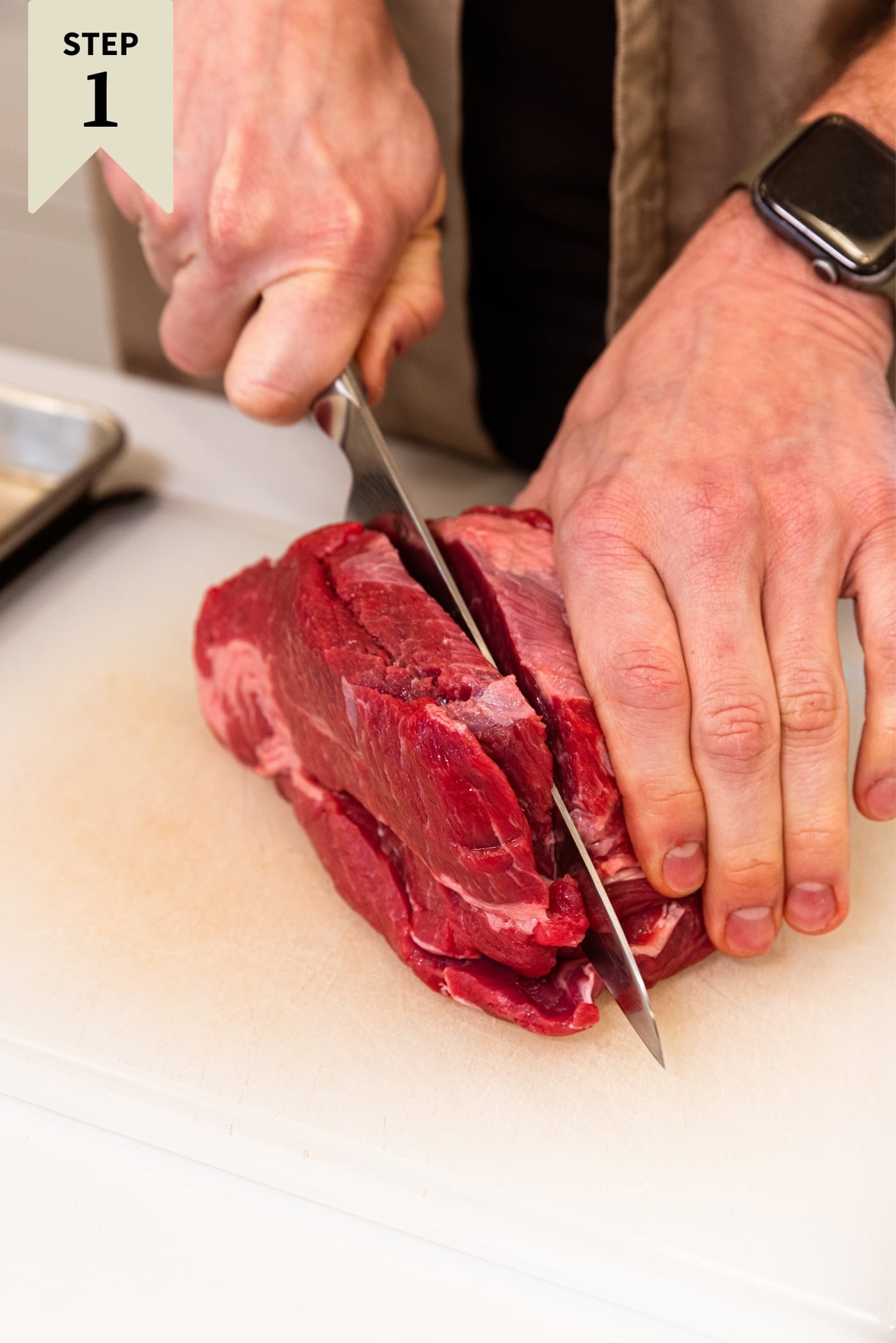 Hand holding raw chuck roast slicing off a piece of a white cutting board.