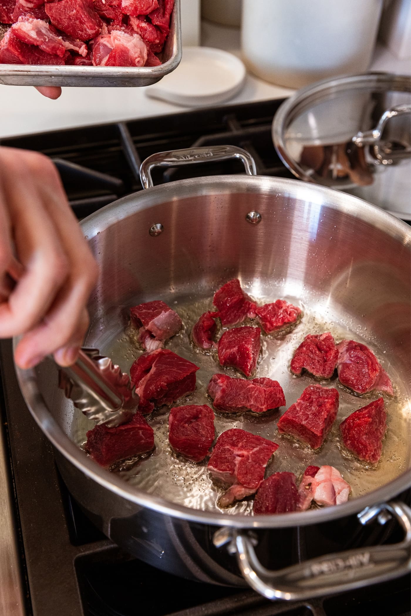 Hand using tongs to place raw pieces of chuck roast in the bottom of a large metal stockpot sitting on stovetop with oil in bottom of pan.