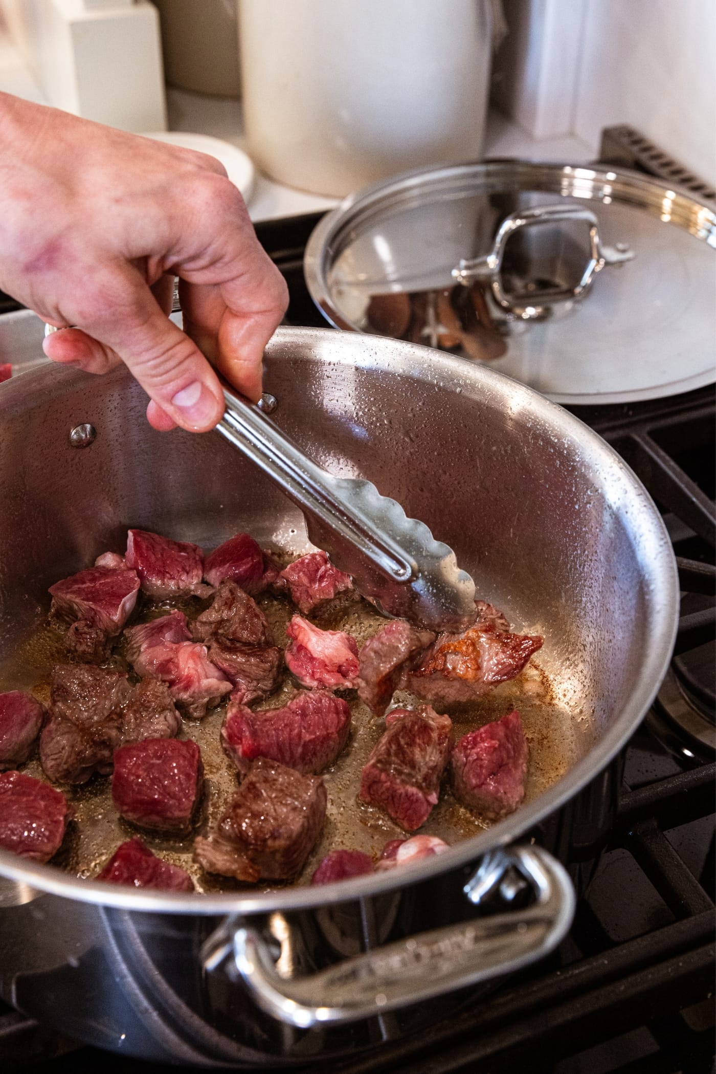 Hand using tongs to turn over half-cooked pieces of chuck roast in metal stockpot sitting on the stovetop cooking the meat.