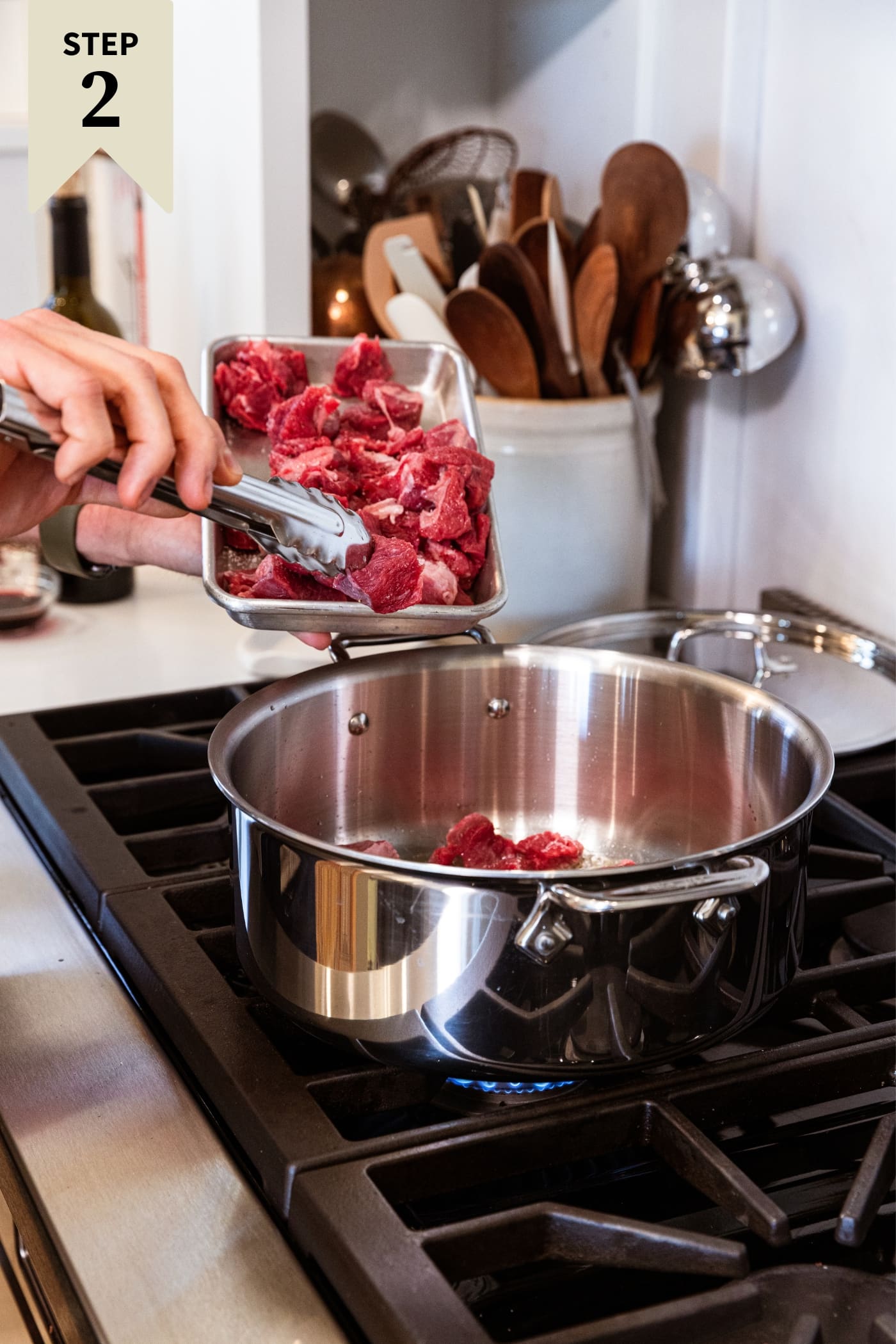 Large metal stockpot on stovetop with hand using tongs to place pieces of raw chuck roast in pot.