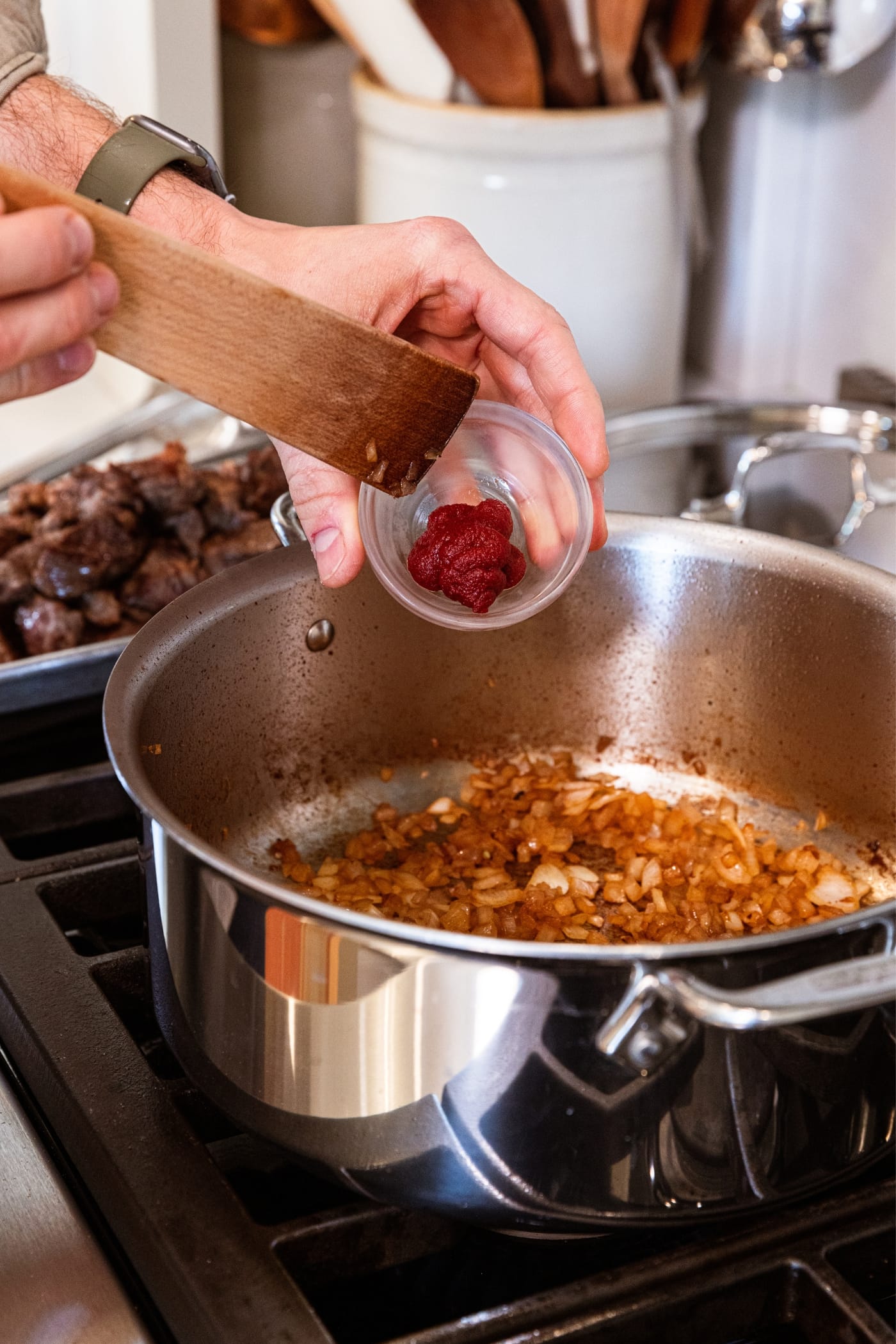 Hand using wooden spatula to scrape out tomato paste from small glass bowl into a large metal stockpot filled with already browned onion pieces.