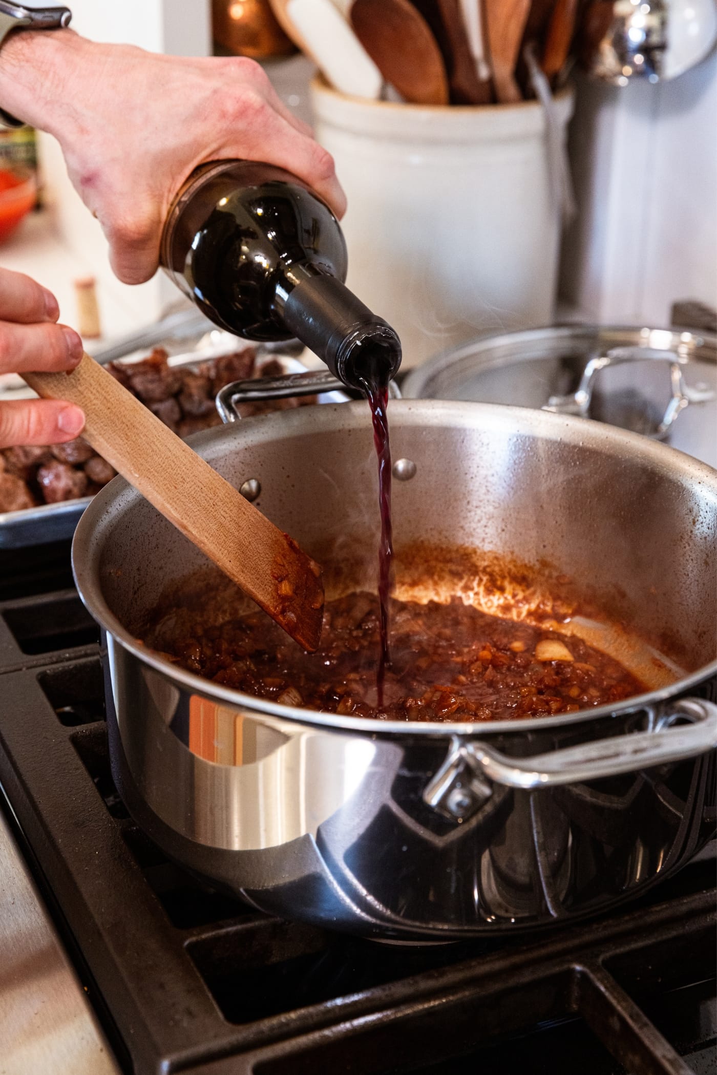 Hand pouring red wine into a large metal stockpot on the stove with diced and browned onions already in the pan.