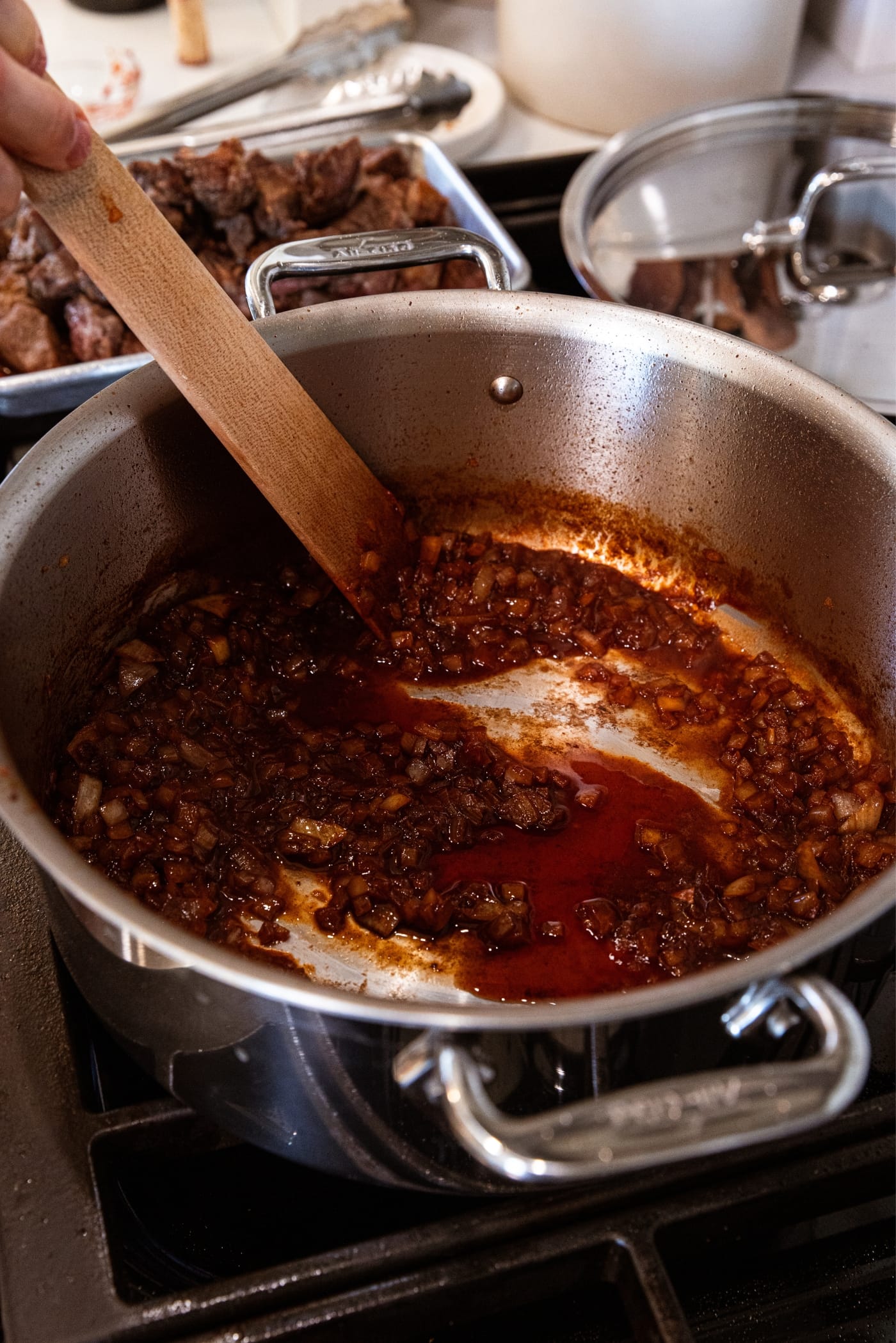 Hand using wooden spatula to mix together red wine and tomato paste with cooked and diced onion mixture in large stockpot on stovetop.