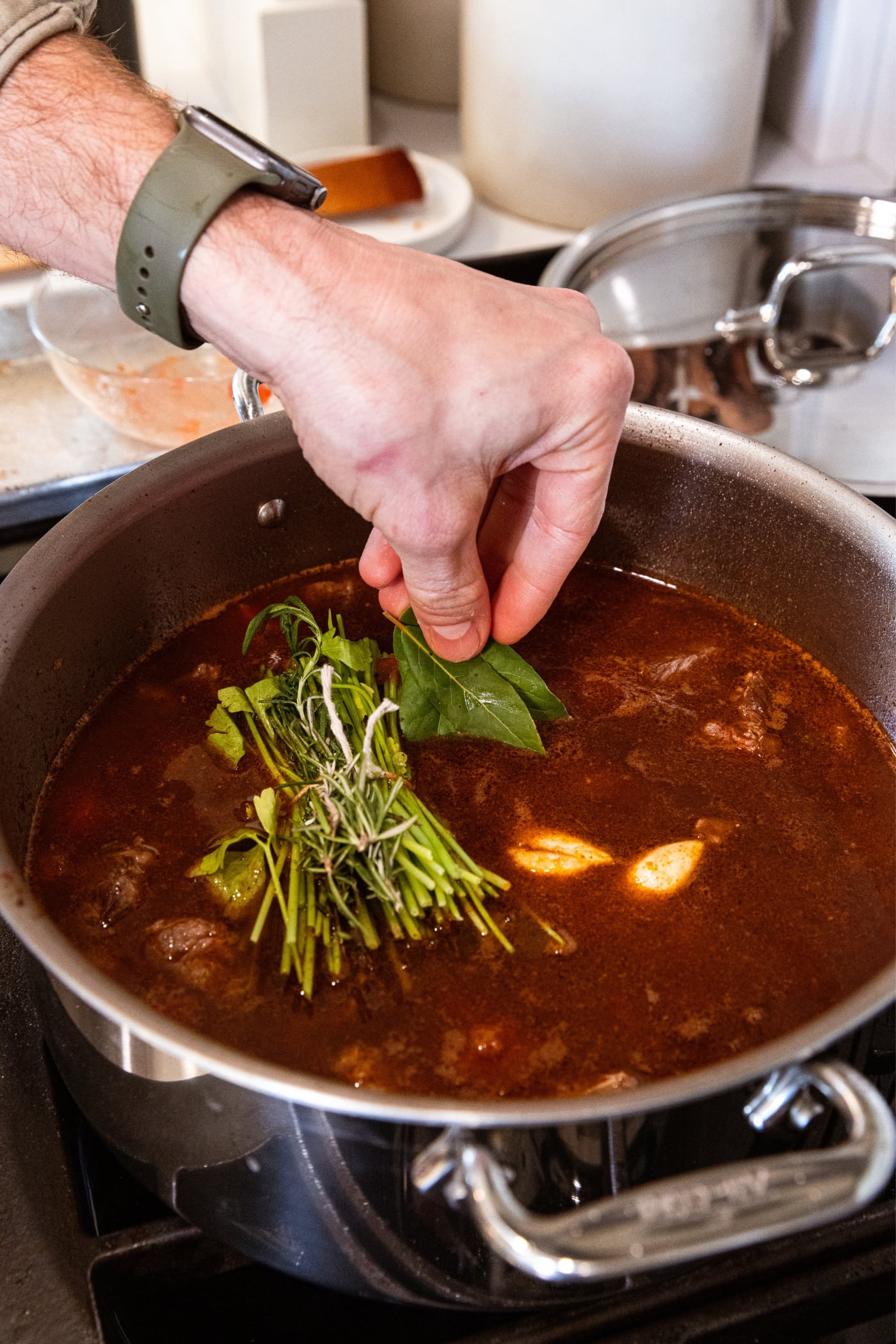 Hand placing packet of herbs into a stockpot filled with cooking soup with garlic cloves floating on the surface along with pieces of browned meat.