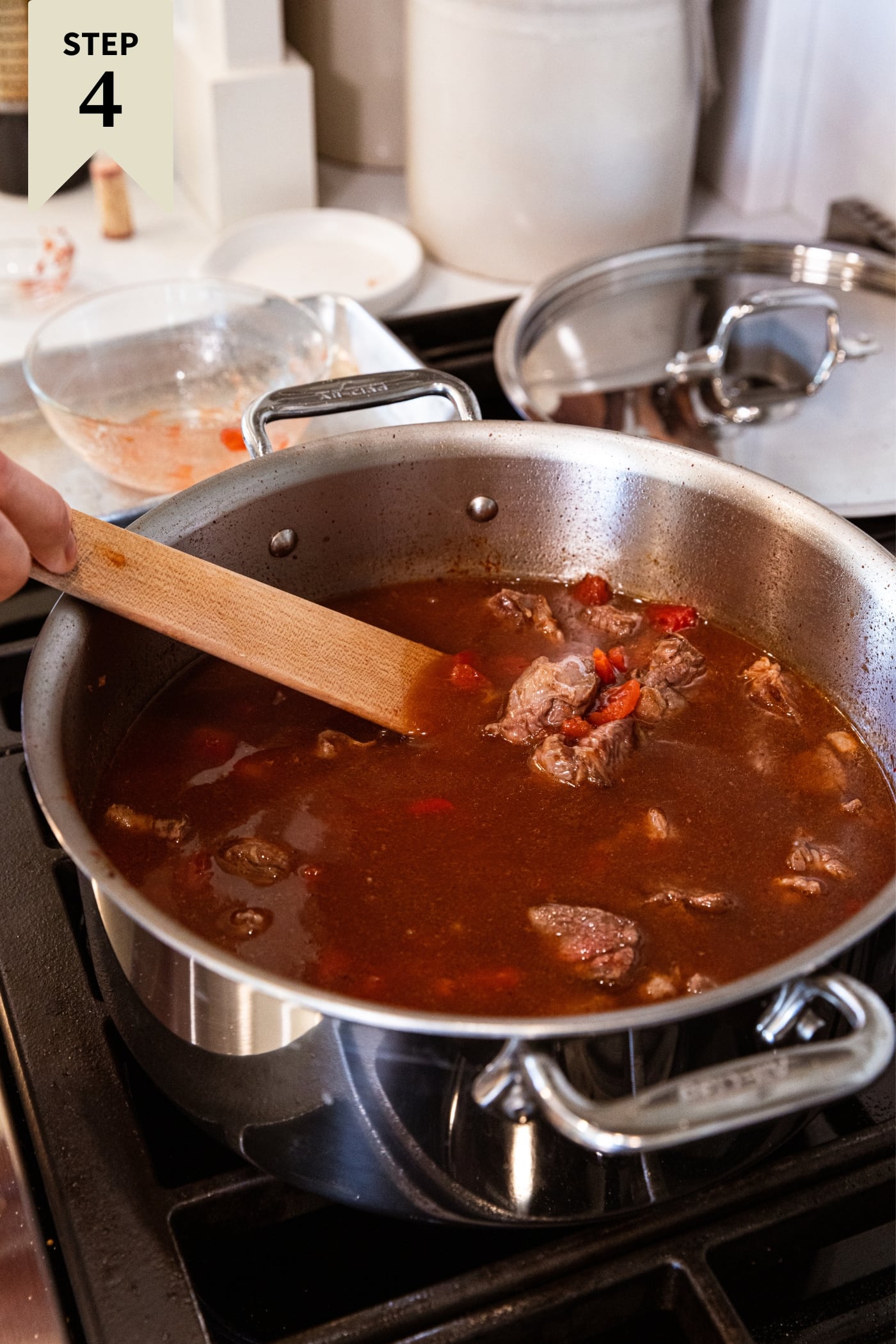 Hand holding wooden spatula mixing together beef with beef stock in large metal stockpot on stovetop with pieces of tomato floating on the surface.